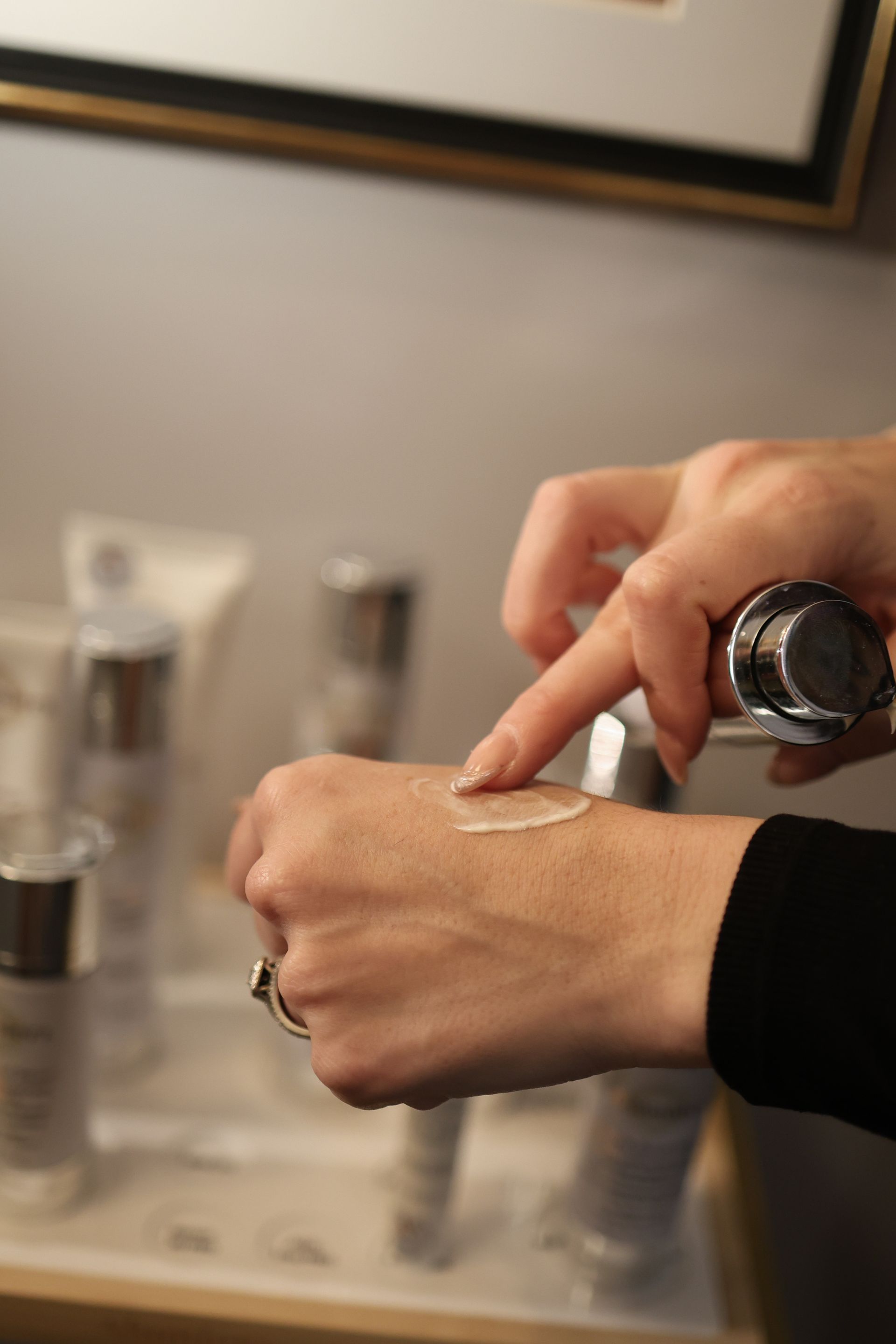 Person applying cream from a silver pump bottle onto their hand, with several cosmetic products in the background.