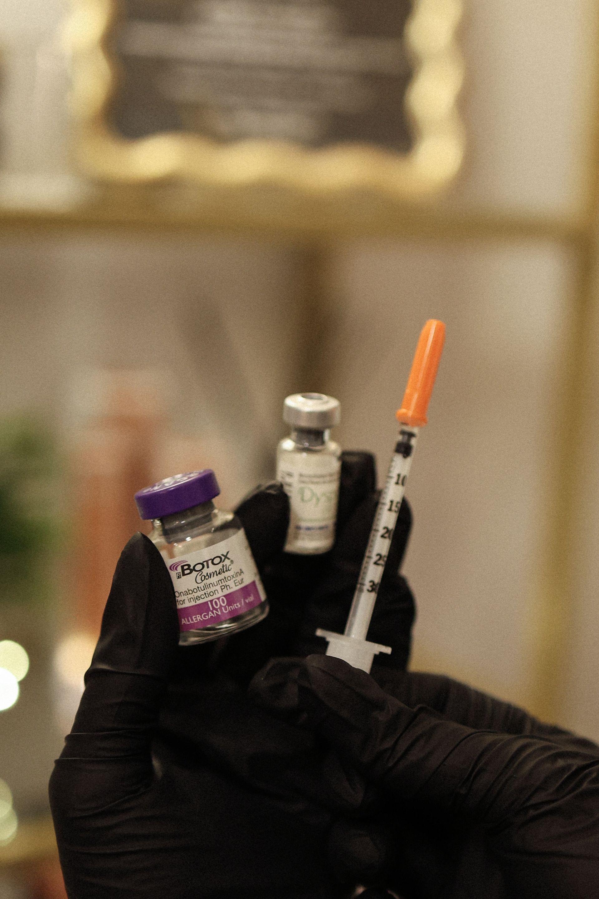 Gloved hands holding vials and a syringe. One vial is purple, another silver. Syringe has an orange cap.