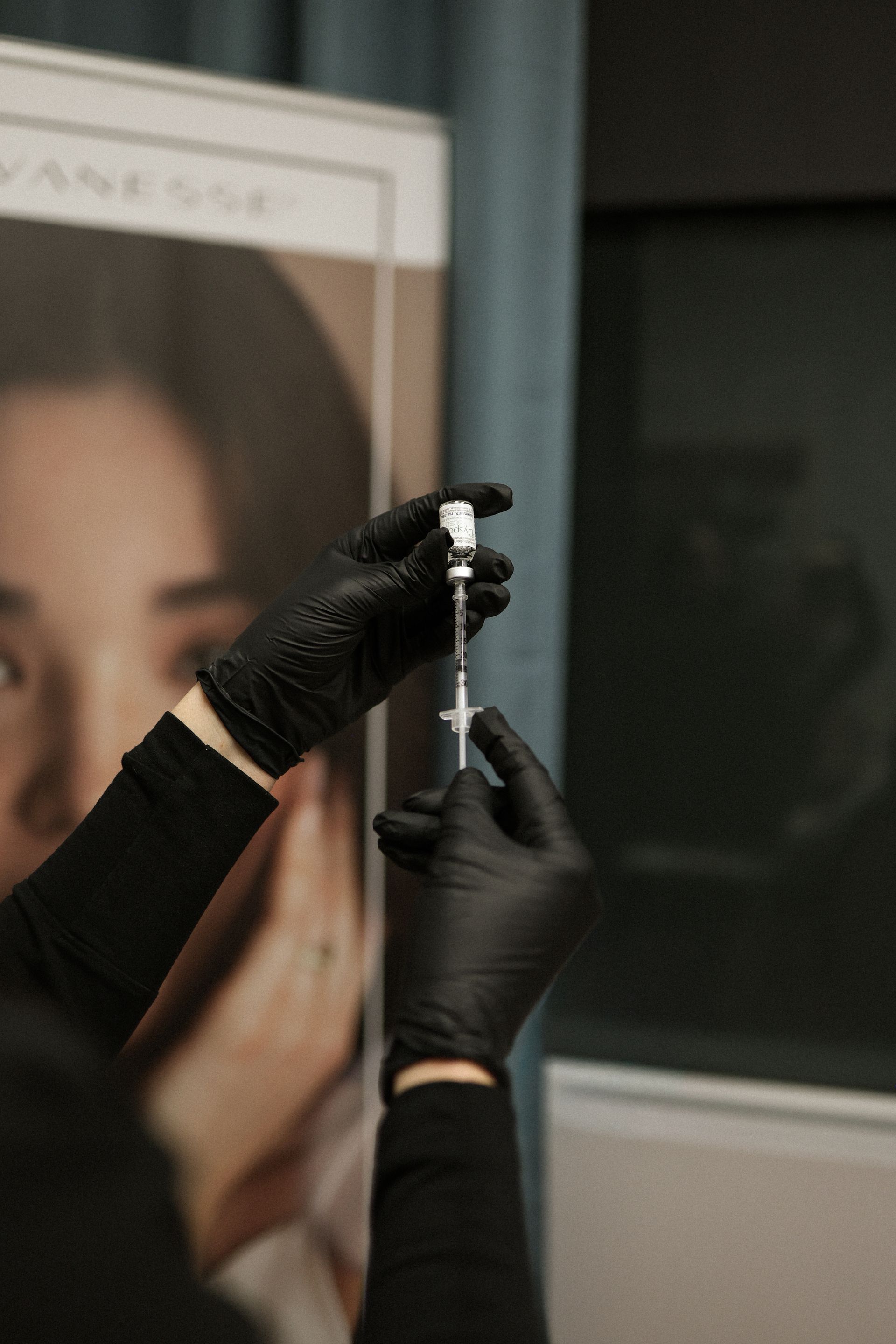 Hands in black gloves fill a syringe from a vial. Background includes a poster and a window.