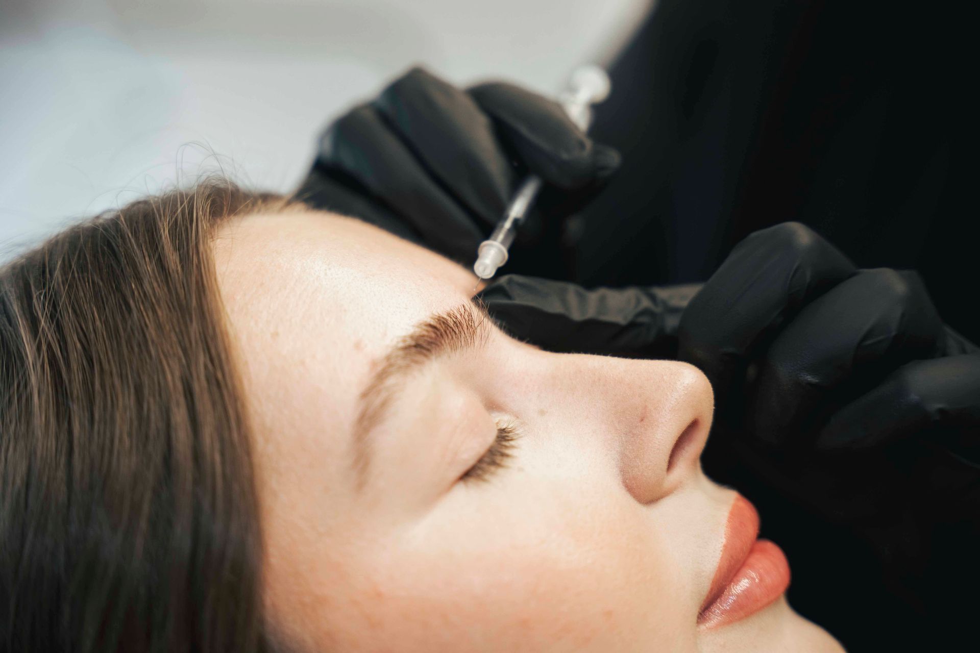 Woman receiving a cosmetic injection in her eyebrow area, administered by someone wearing black gloves.