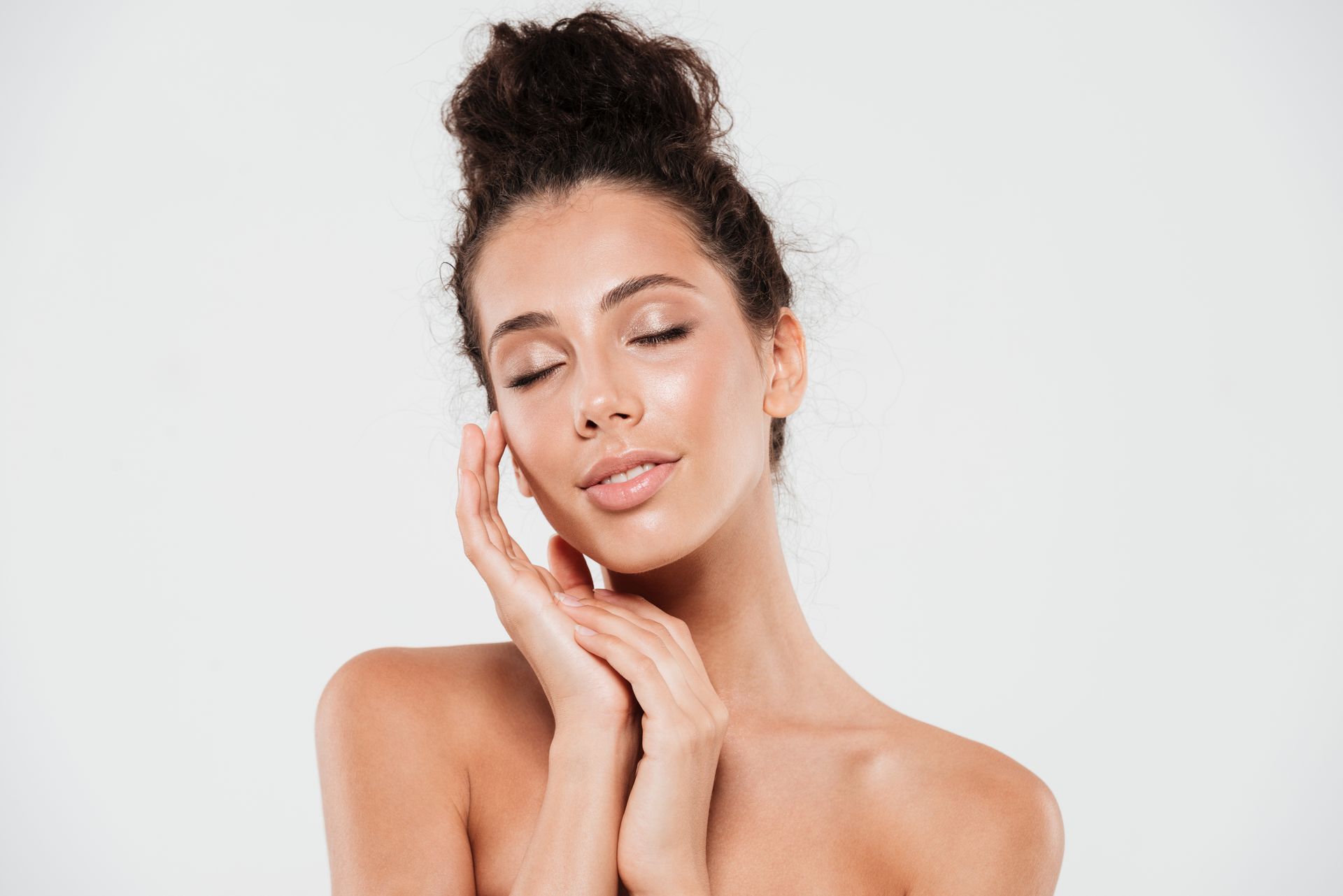 Woman with eyes closed, hands on face, smiling; light skin, bun hairstyle, neutral background.