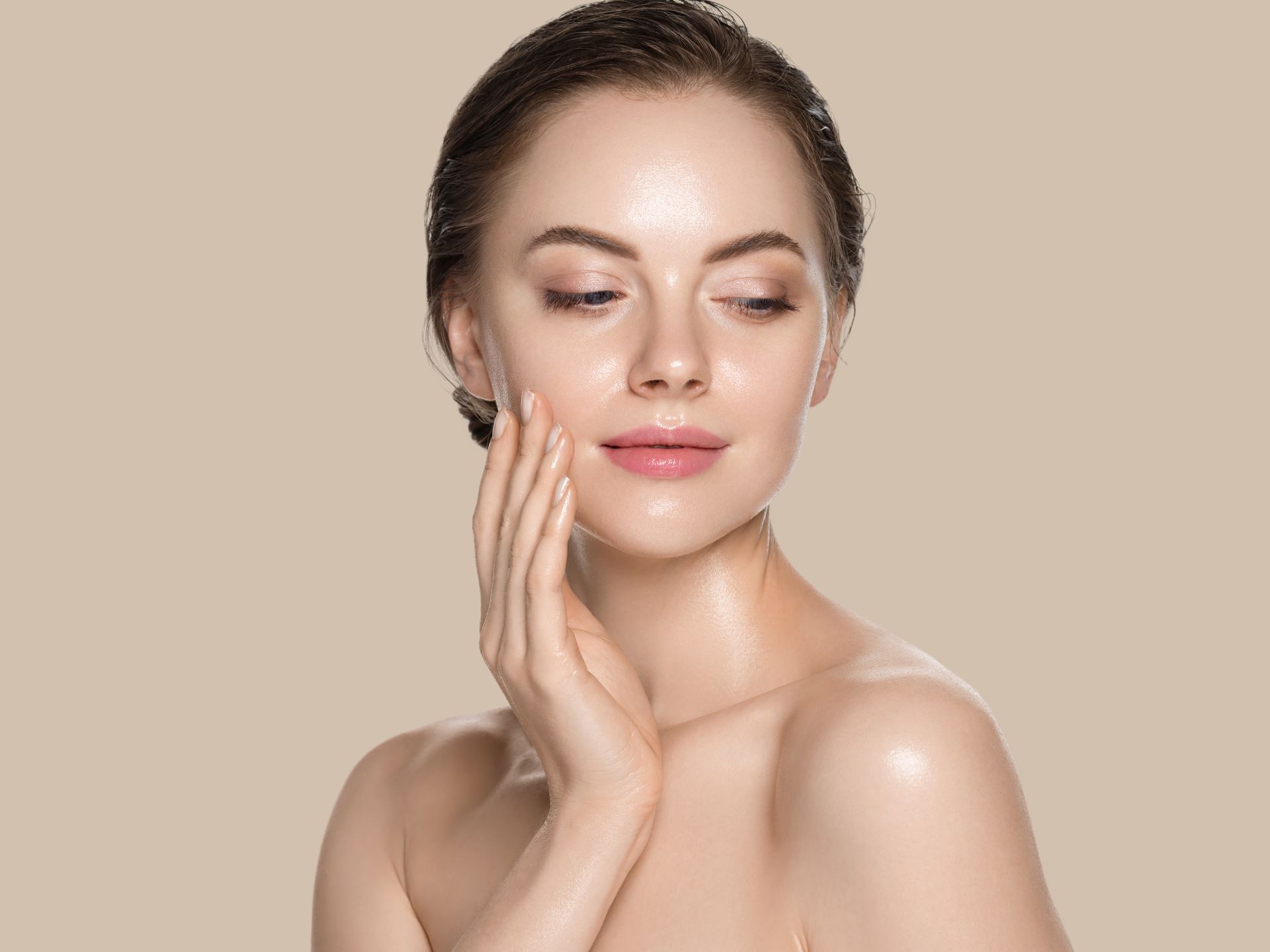 Woman with glowing skin, touching her face, nude shoulders, soft lighting.