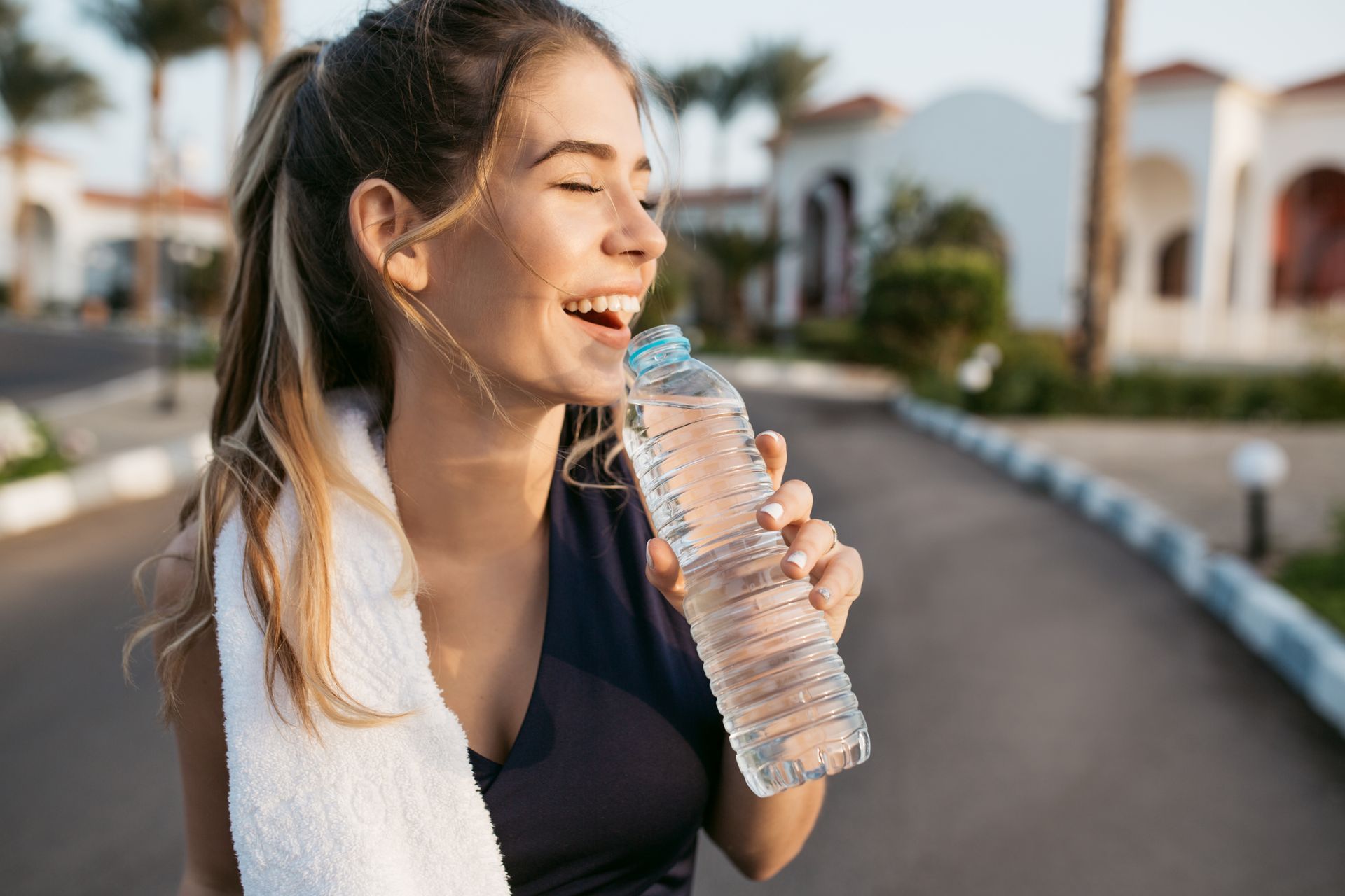 Woman in athletic wear drinks water, smiling, towel over shoulder. Outdoors, sunny.