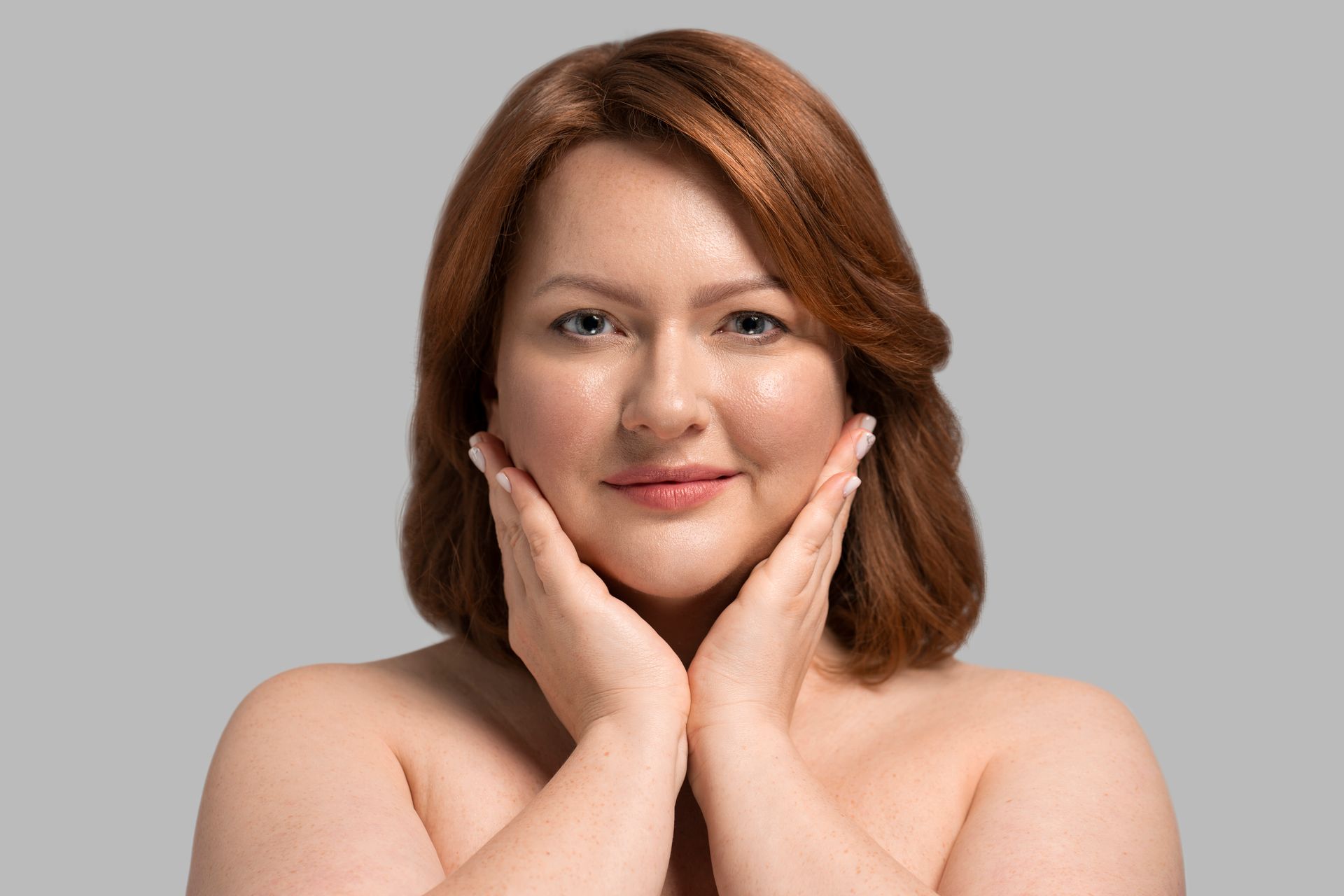 Woman with auburn hair, holding hands to cheeks, smiles softly against a gray background.