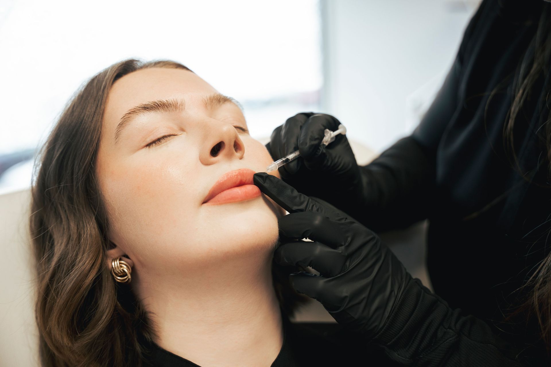 A woman receives lip filler injections from a person wearing black gloves in a clinic setting.
