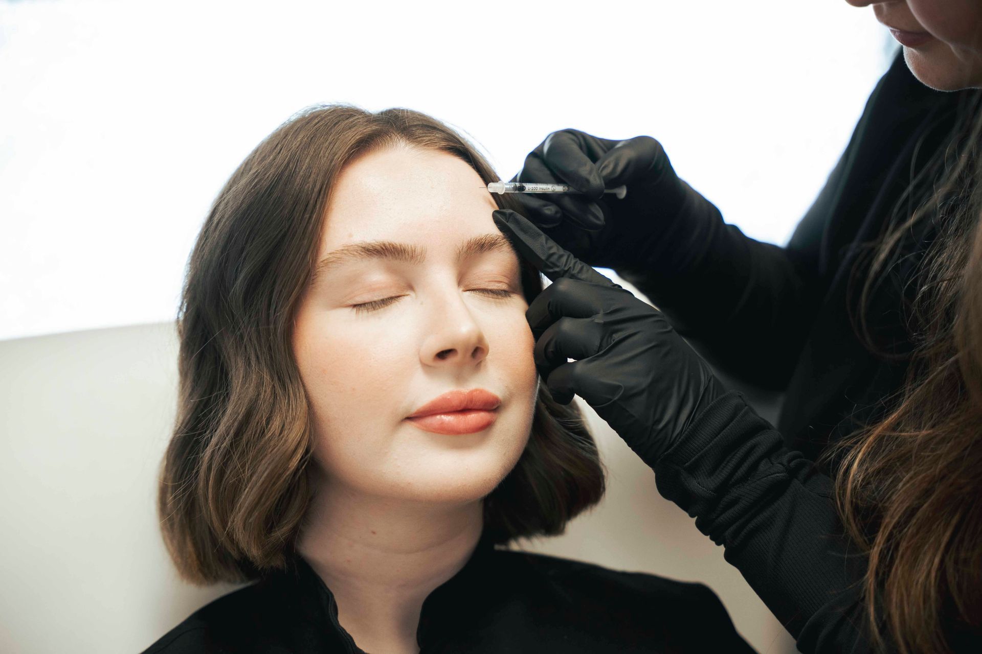 Woman receiving a cosmetic injection near the eyebrow; eyes closed, black gloves.