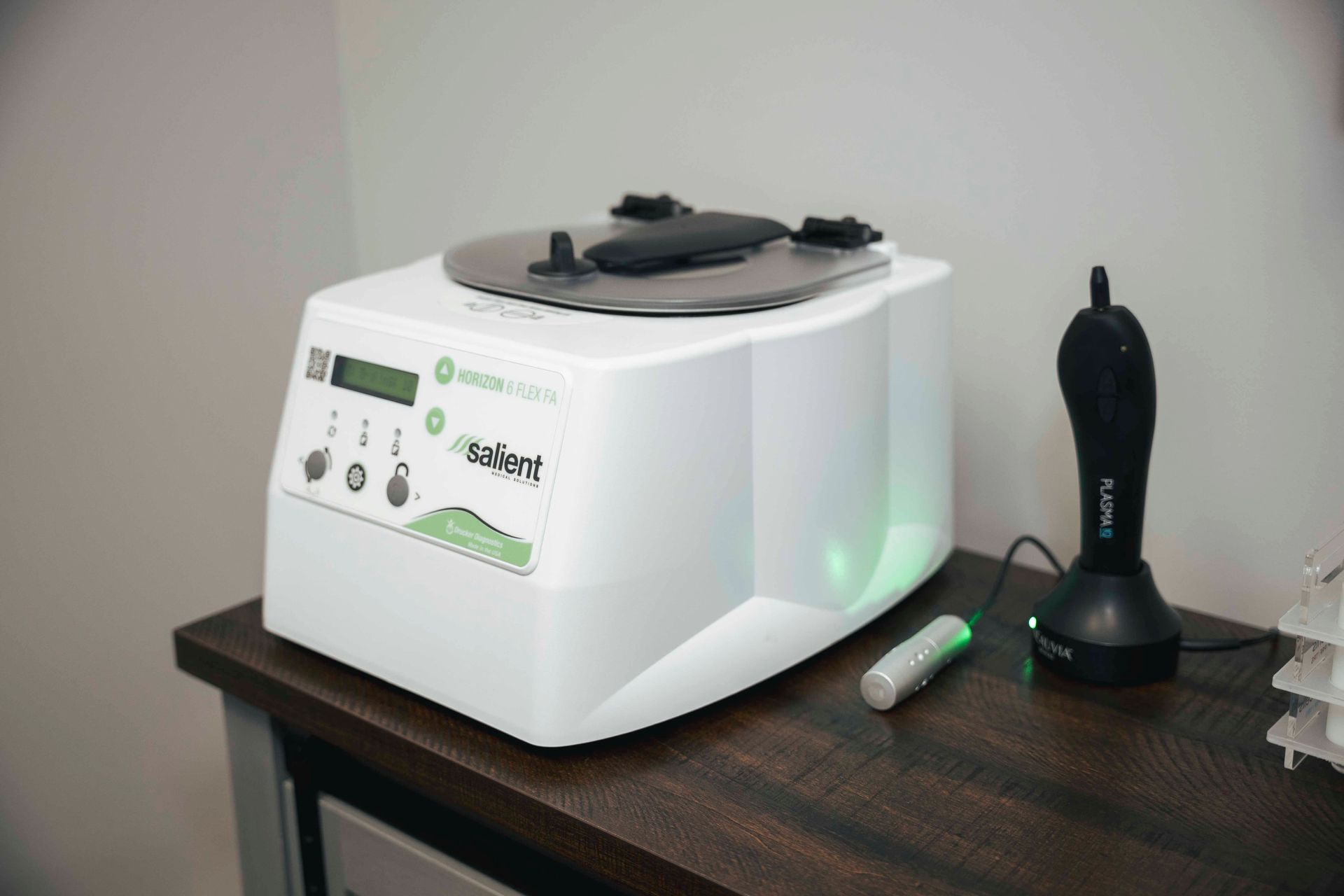 White medical device on a dark desk with a green LED light. A black handheld device rests beside it.