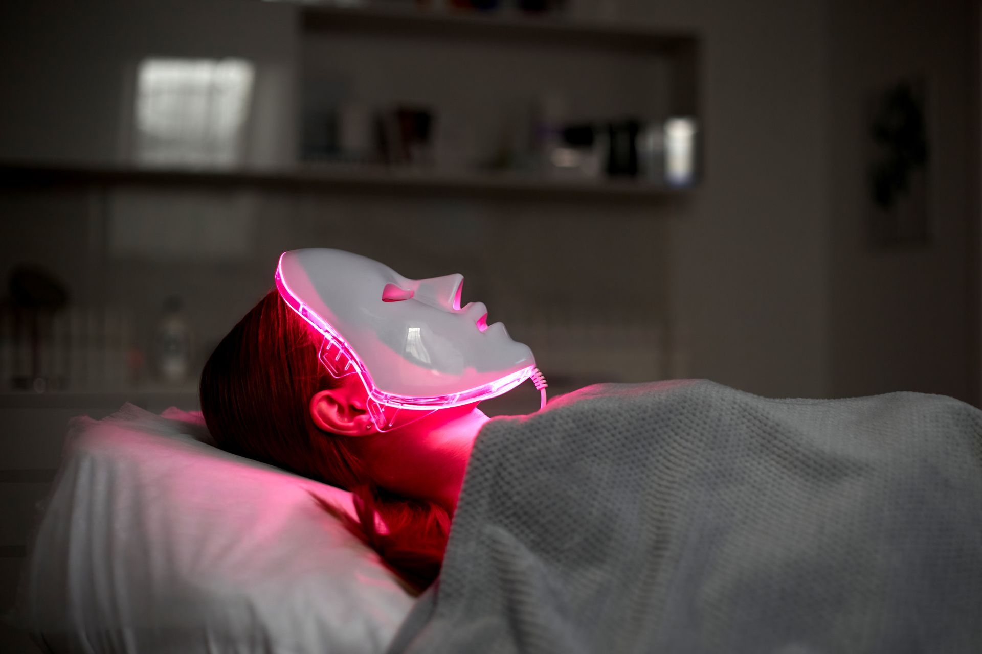 Person wearing a red-light therapy mask, reclined on a spa bed in a dimly lit room.