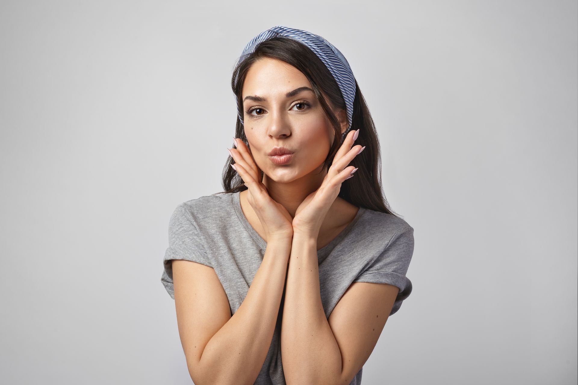 Woman with dark hair wearing a blue headband, gray shirt, pouting lips, and hands framing her face, on a gray background.