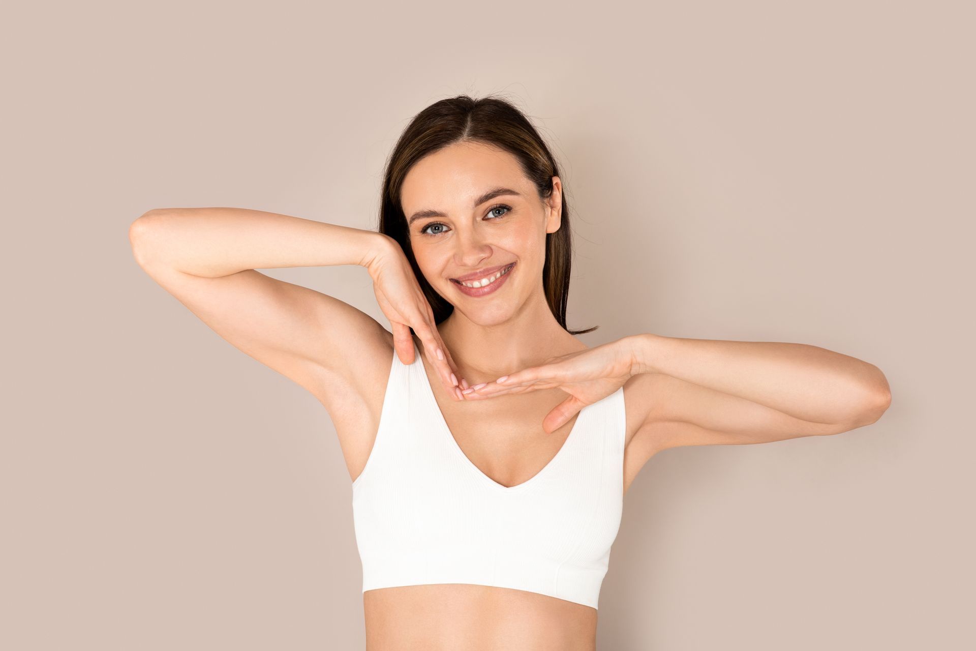Woman in white sports bra smiles, touching face and chest, neutral background.