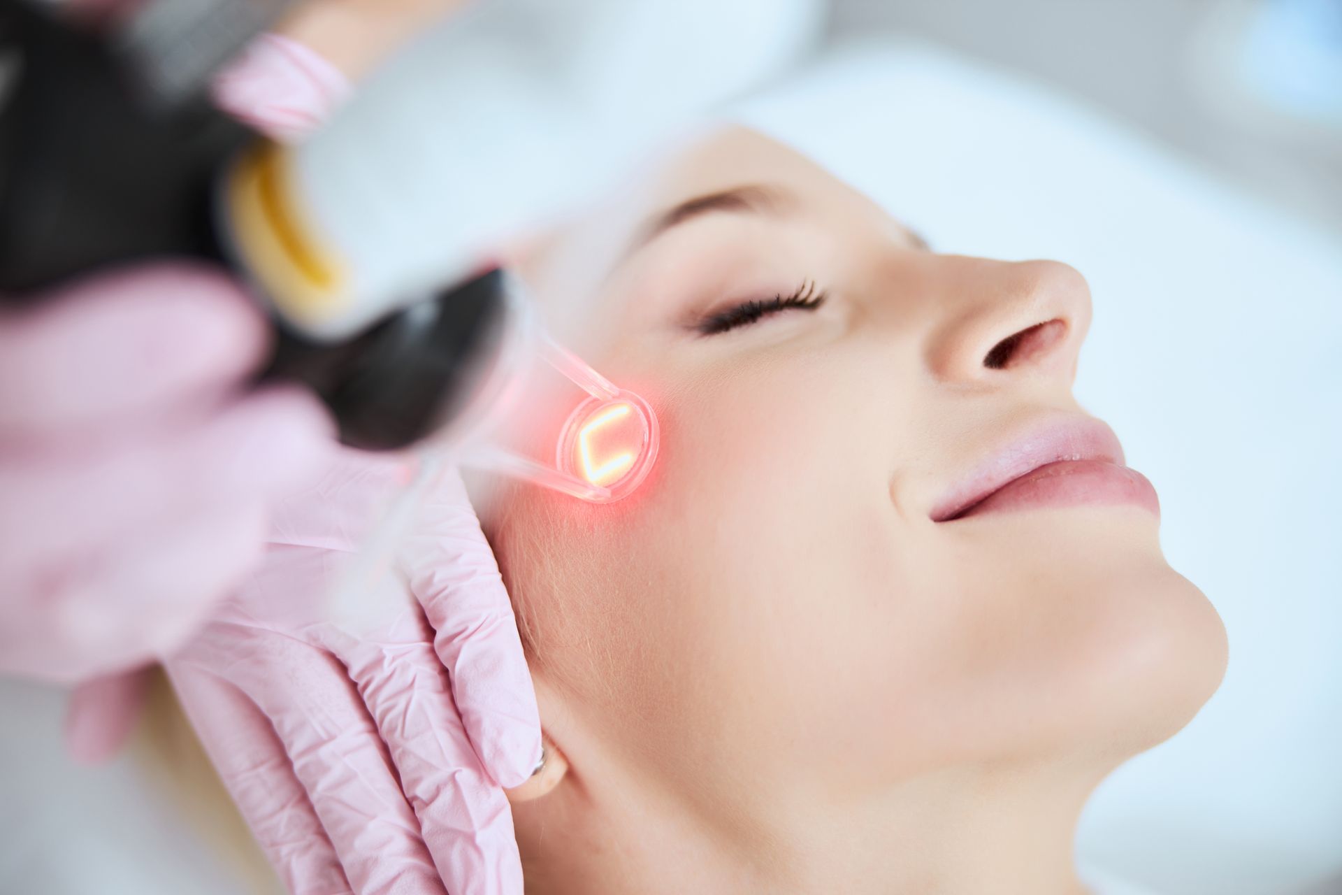 Woman receiving facial laser treatment; red light on cheek, eyes closed. Pink gloves.