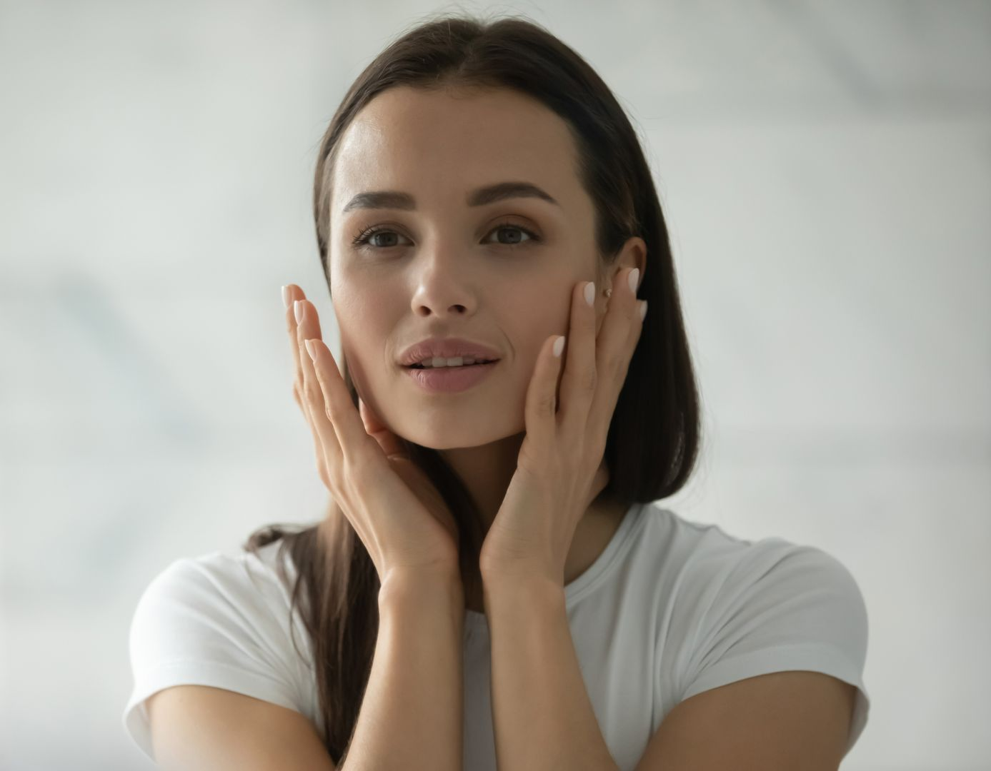 Woman looking in a mirror, touching her cheeks, white t-shirt, marble wall backdrop.