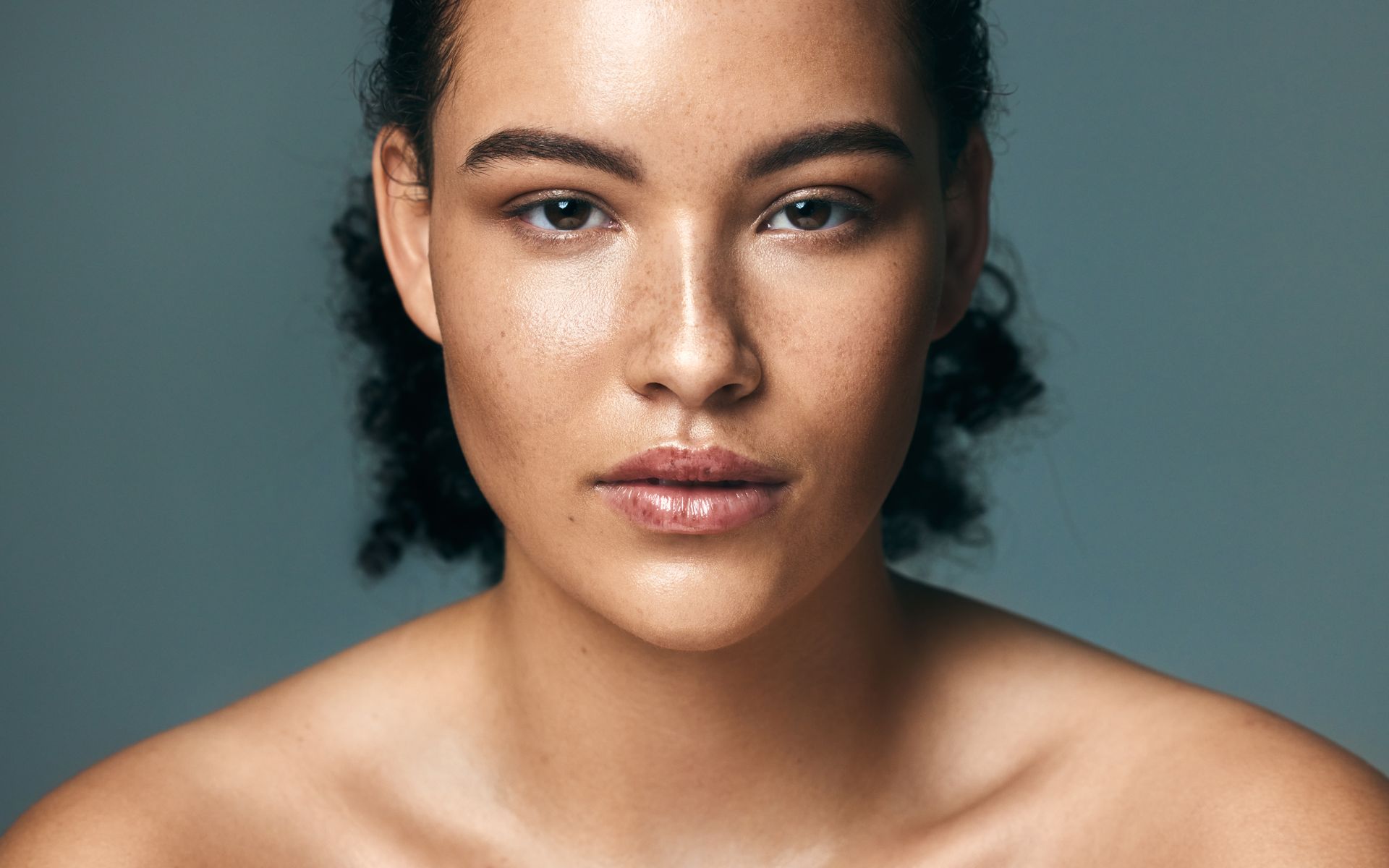 Woman in white tank top smiles, touches face, brown hair, neutral background.