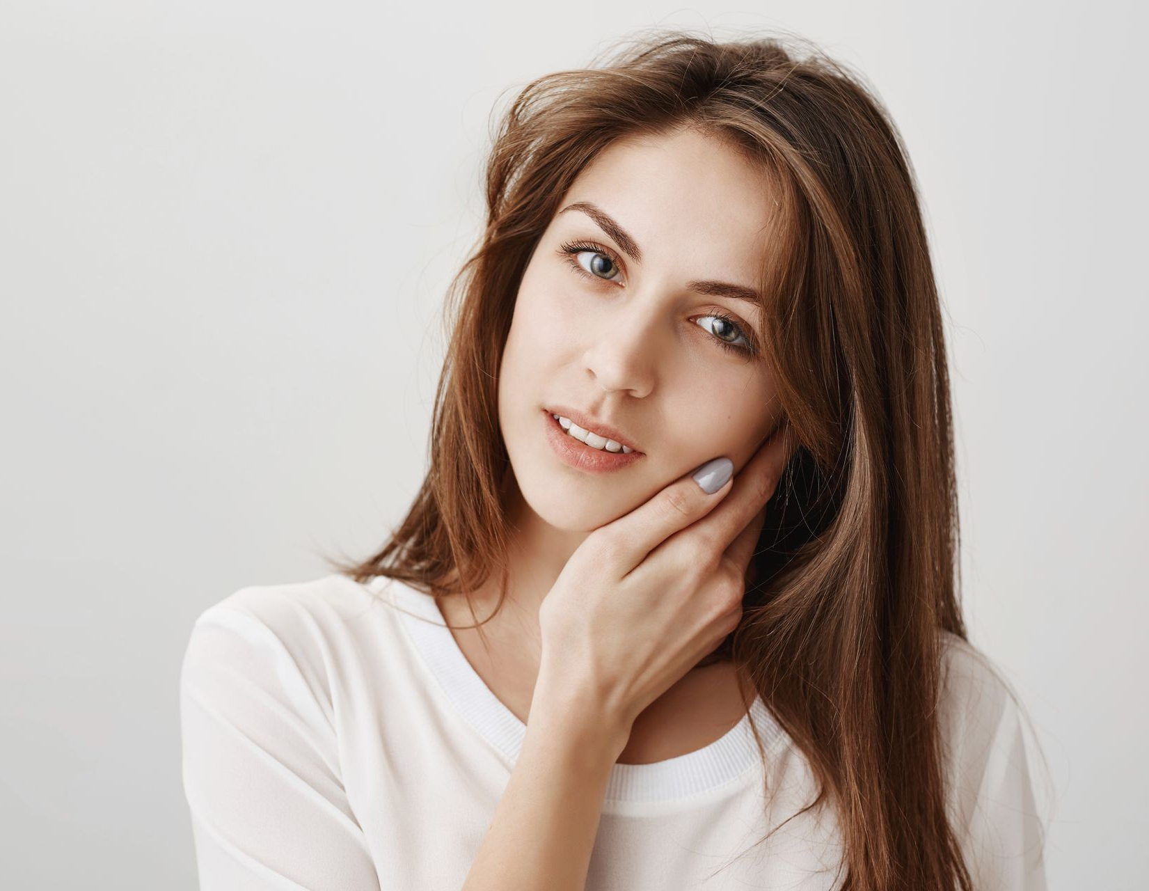 A person with long brown hair wearing a white shirt, looking at the camera while resting their cheek on their hand.