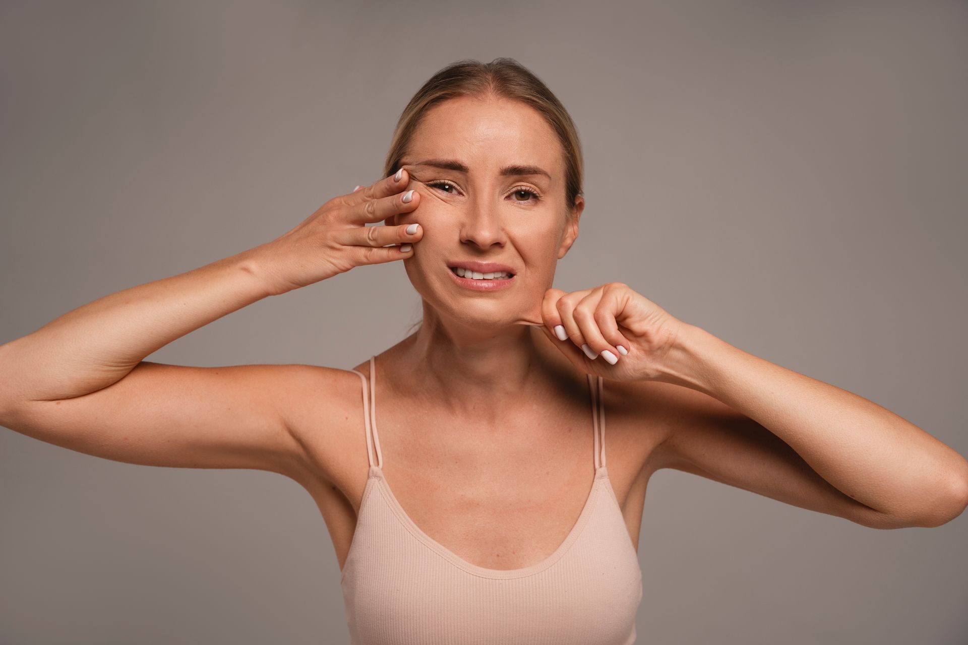 Woman pulling at face and neck skin, showing signs of aging, in front of gray background.