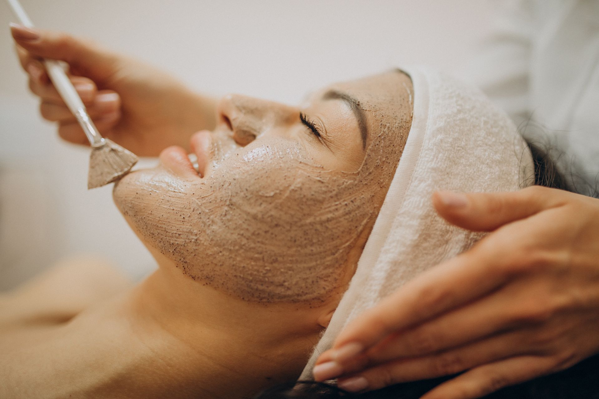 Woman receiving a facial treatment at a spa, with a face mask being applied by hand.