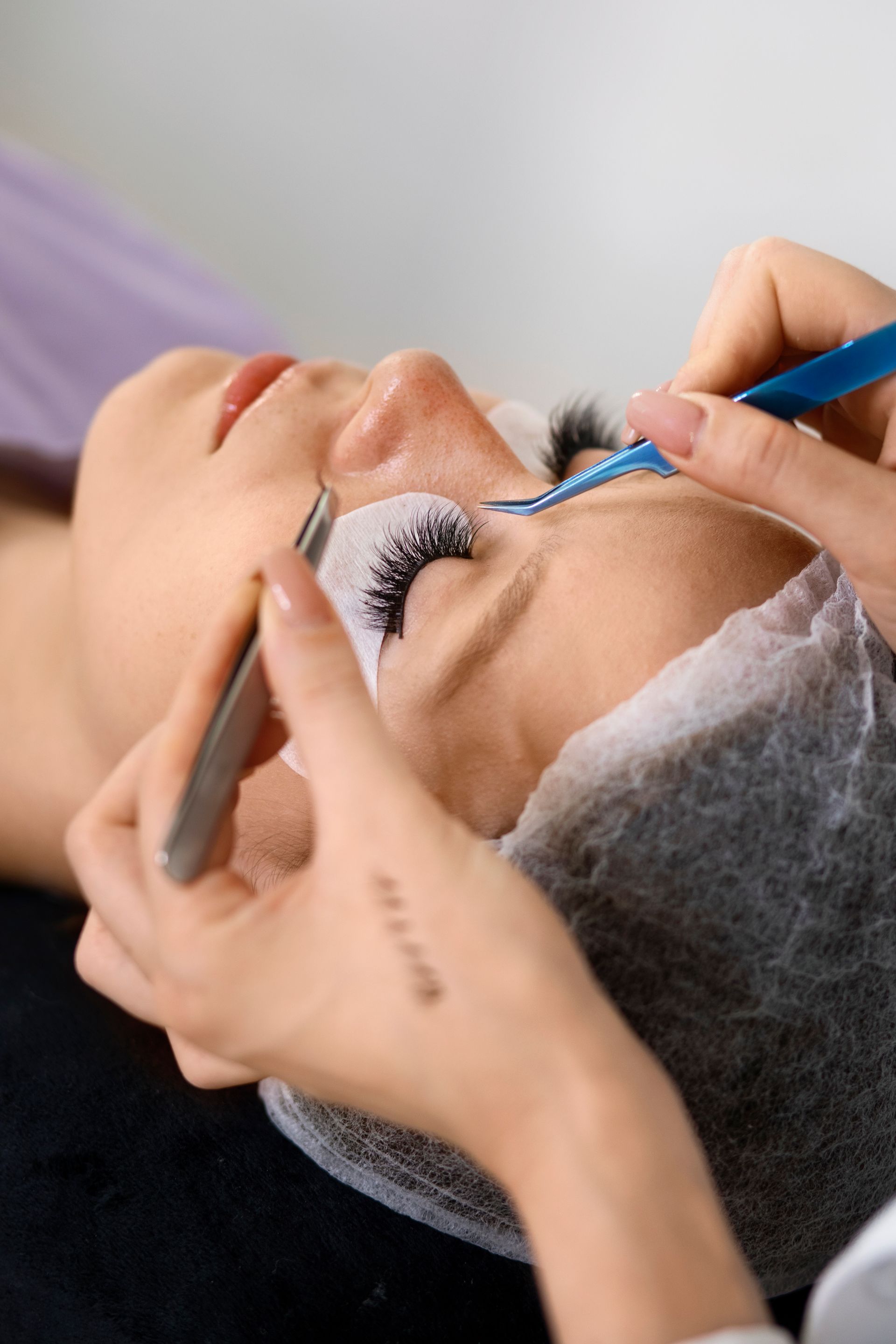 Woman receiving eyelash extensions; technician applying with tweezers.
