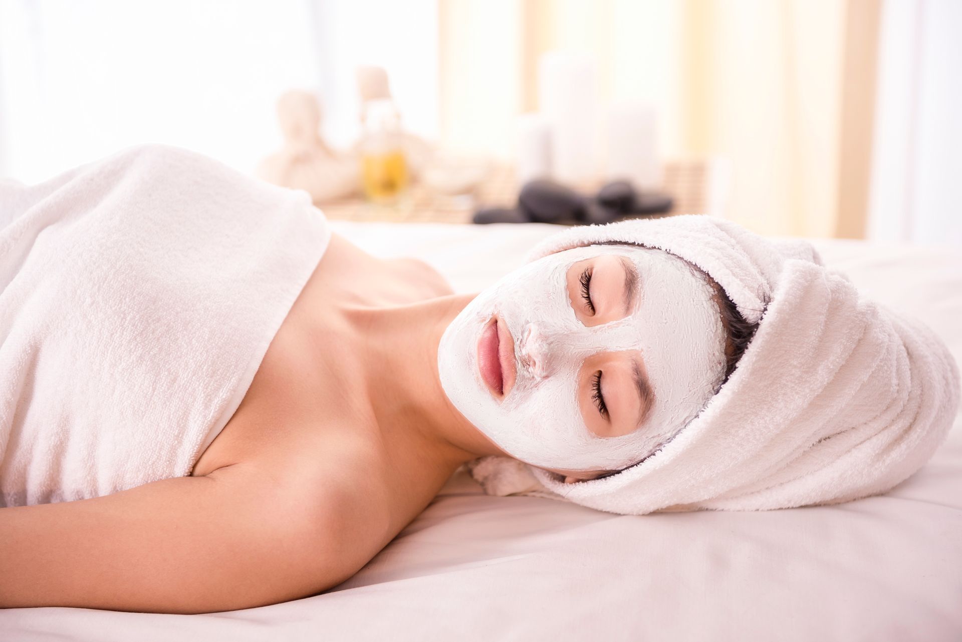 Woman with face mask and towel on head, relaxing on spa bed.
