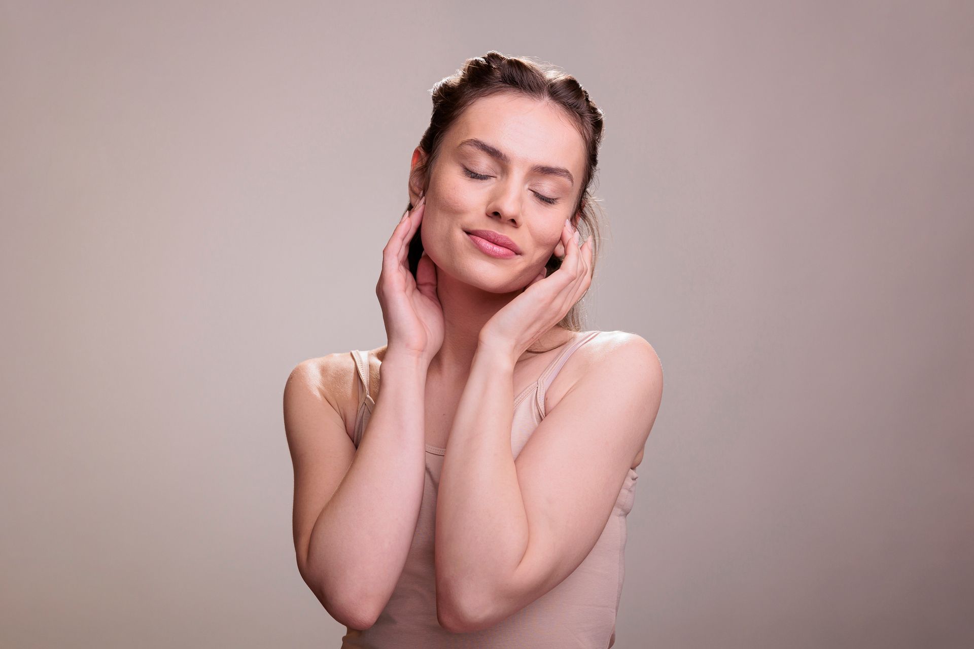 Woman with eyes closed, touching her face gently. Brown hair up, neutral-toned top, soft background.