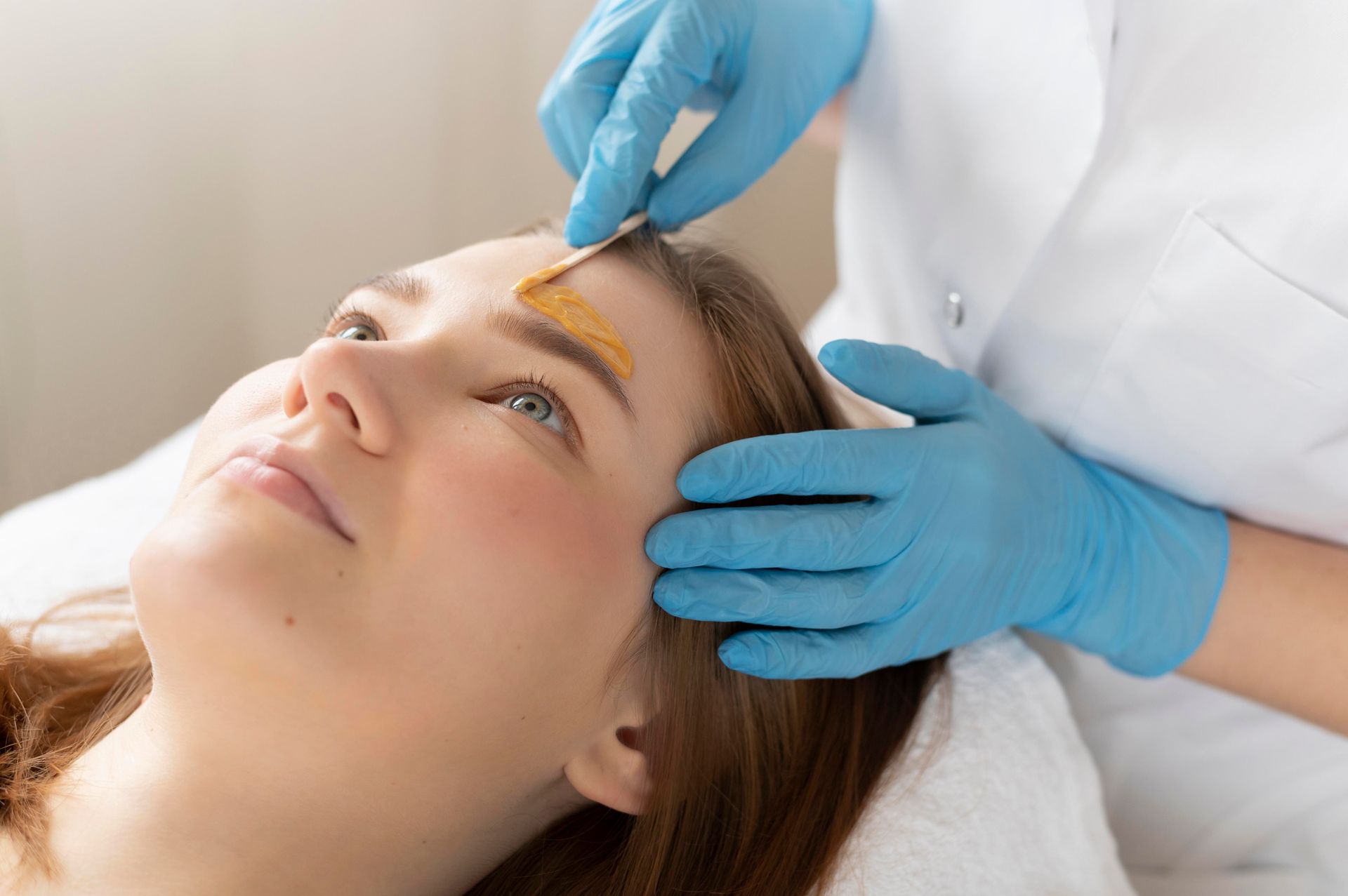 Woman getting eyebrow waxed at a salon; beautician wearing gloves applies wax with a spatula.