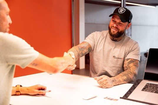 Twee mannen schudden elkaar de hand aan een bureau; een van hen draagt ​​een hoed, tatoeages en glimlacht.