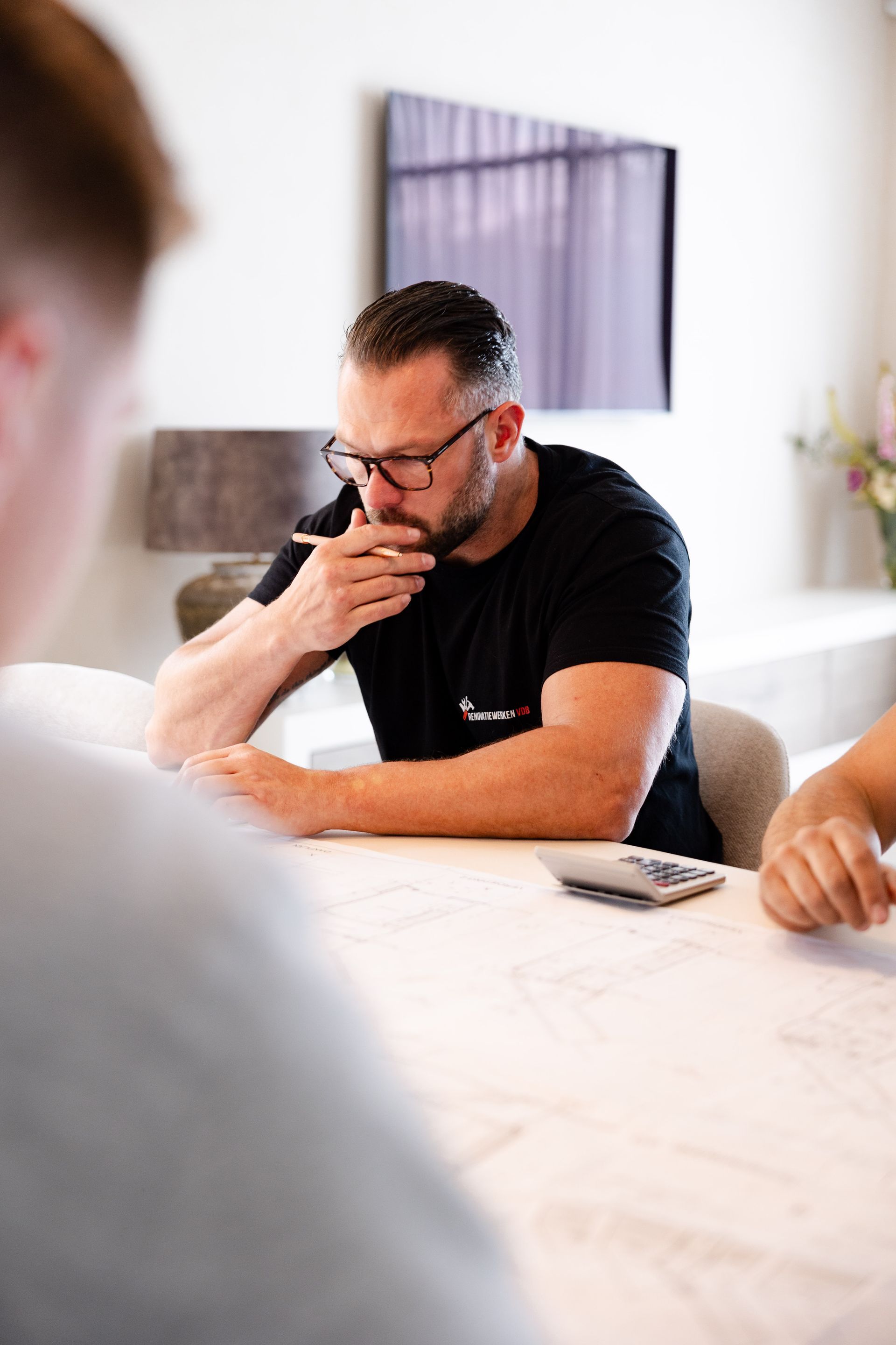 Man met bril, zittend aan tafel, nadenkend kijkend, pen bij zijn mond houdend.