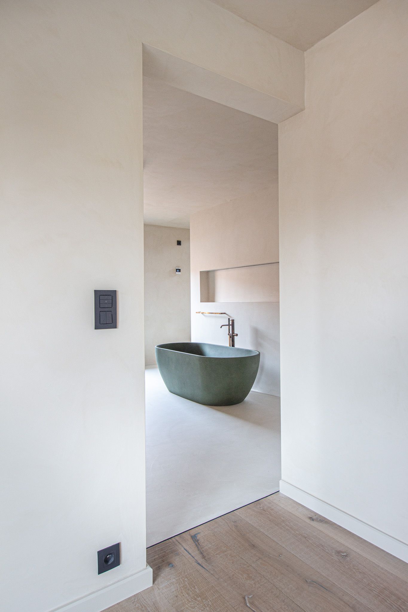 Een smalle doorkijk naar een badkamer met een groen stenen bad. Lichtgekleurde muren en vloer.