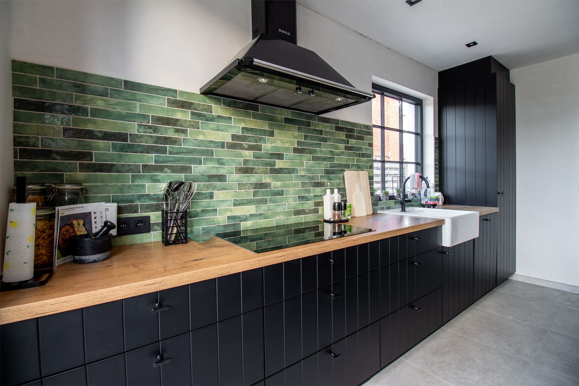 Zwarte keukenkastjes met houten aanrechtbladen, groene tegelachterwand en een zwarte afzuigkap.