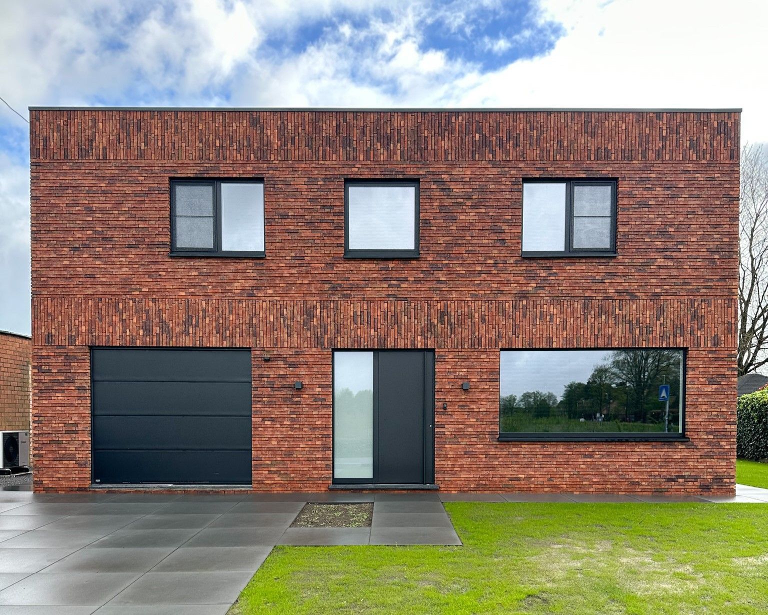 Bakstenen huis met twee verdiepingen, garage, voordeur en ramen. Rode bakstenen, zwarte accenten en een groen gazon.
