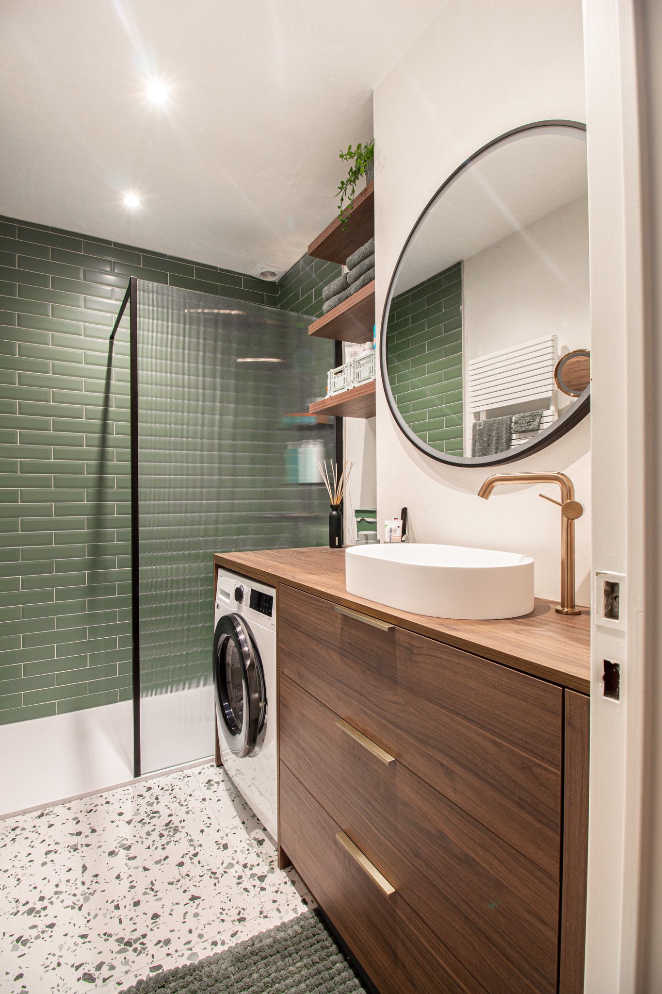 Moderne badkamer met groene tegels, houten wastafel, ronde spiegel en wasmachine/droger.