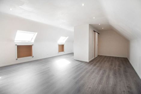 Lege zolderkamer met grijze houtlook vloer, witte muren en kleine ramen met bruine jaloezieën.