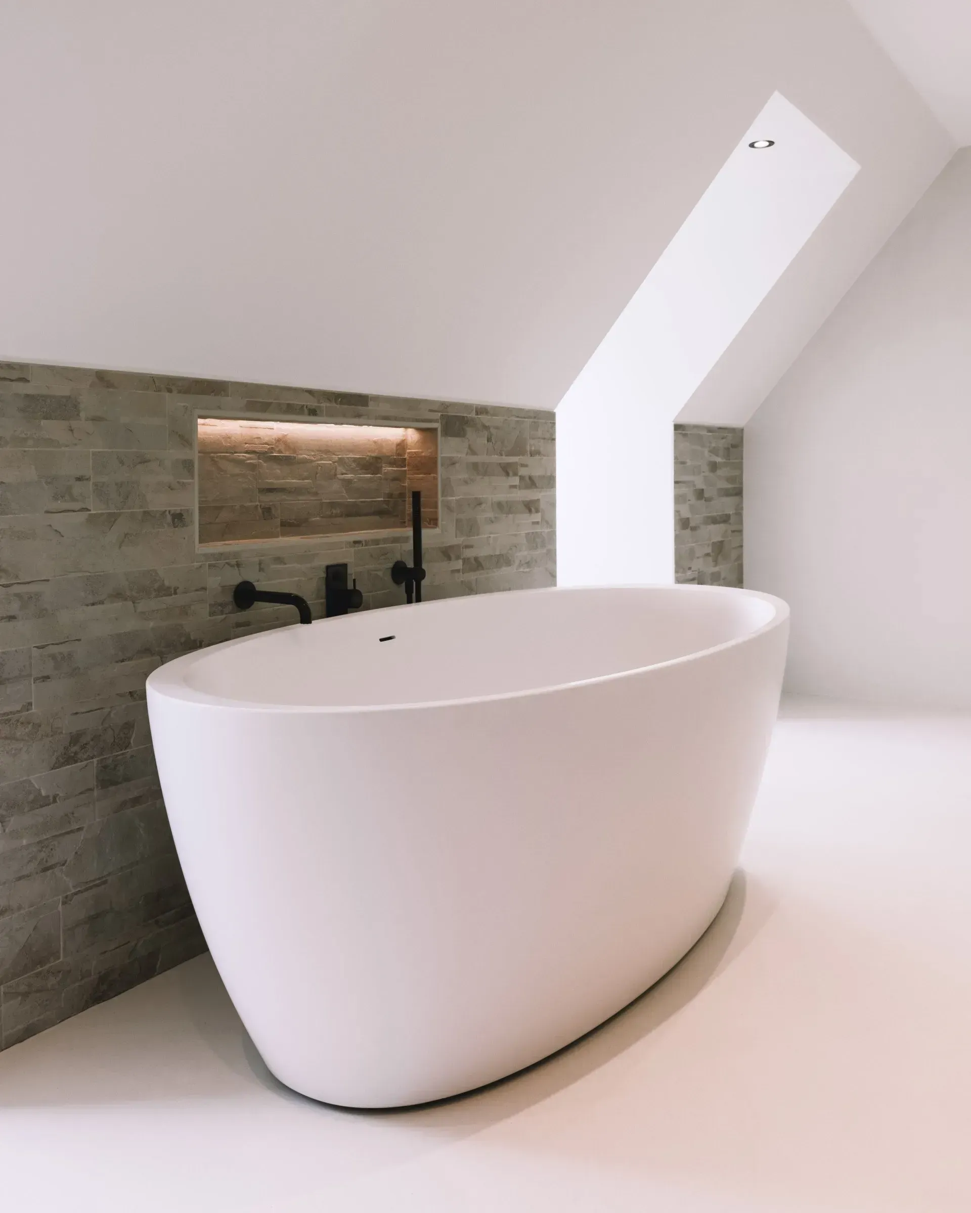 Wit, ovaal bad in een moderne badkamer met een schuin plafond en een verdiepte muurnis.