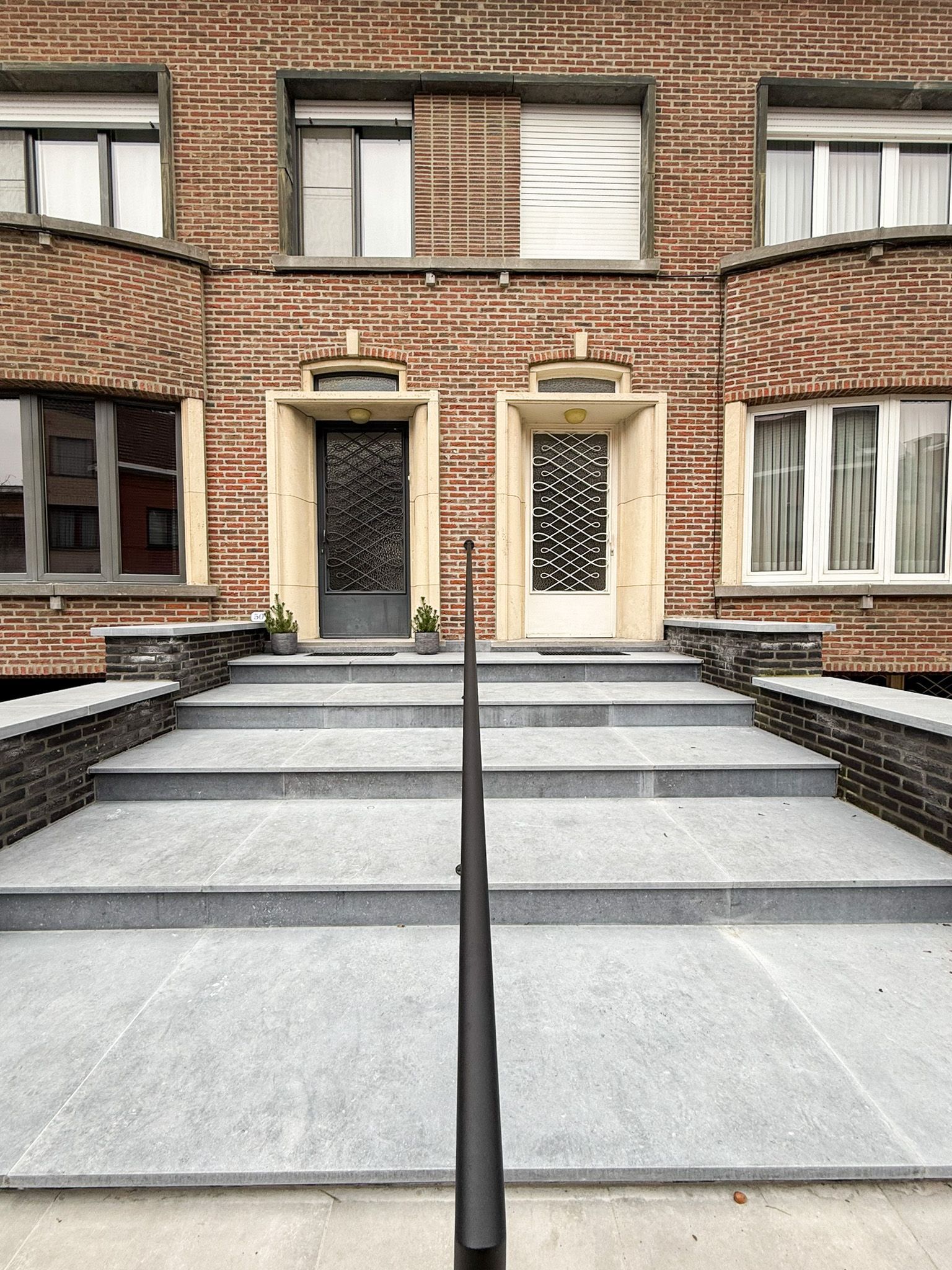 Bakstenen gebouw met twee deuren die toegankelijk zijn via trappen, zwarte leuning op de voorgrond.