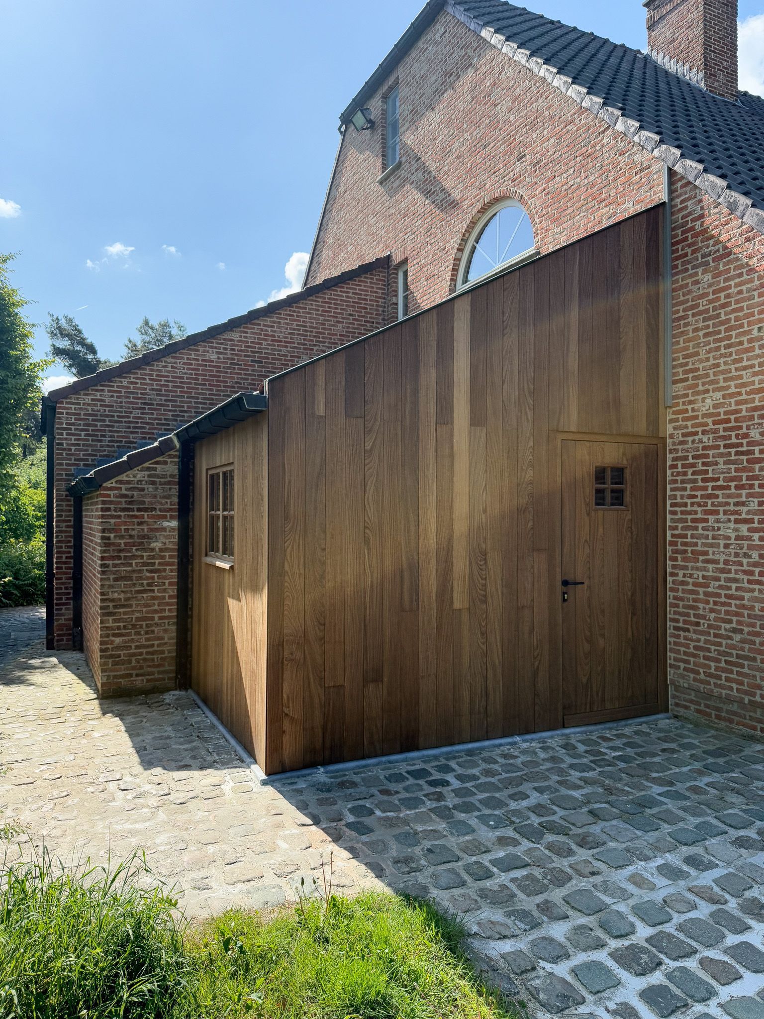 Buitenkant van een gebouw met lambrisering van baksteen en hout. Bovenin bevindt zich een rond raam. Looppad van blauwe kasseien.