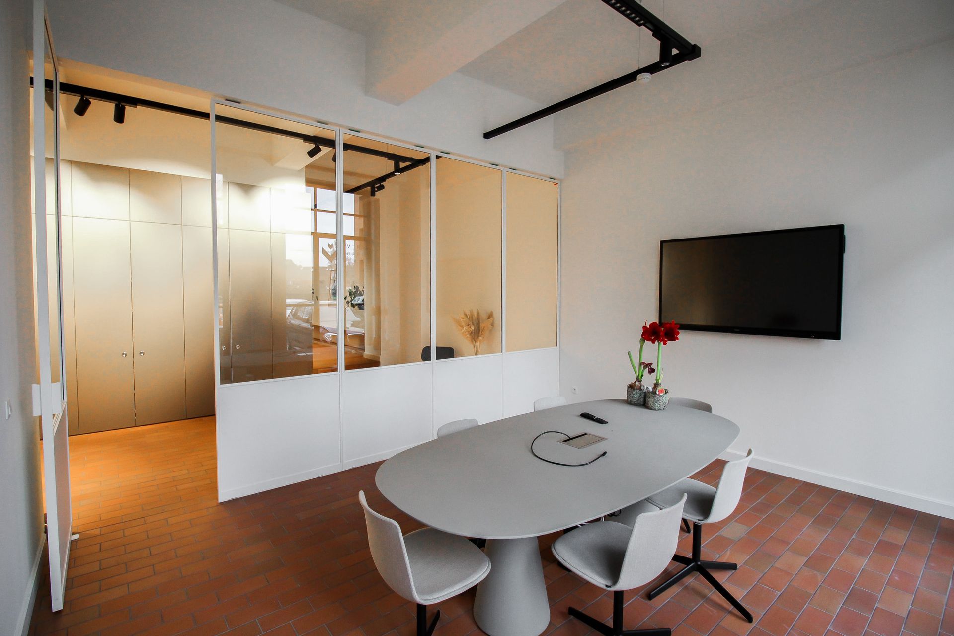 Vergaderruimte met ovale tafel, stoelen en tv. Glas en witte muur. Rode tegelvloer.