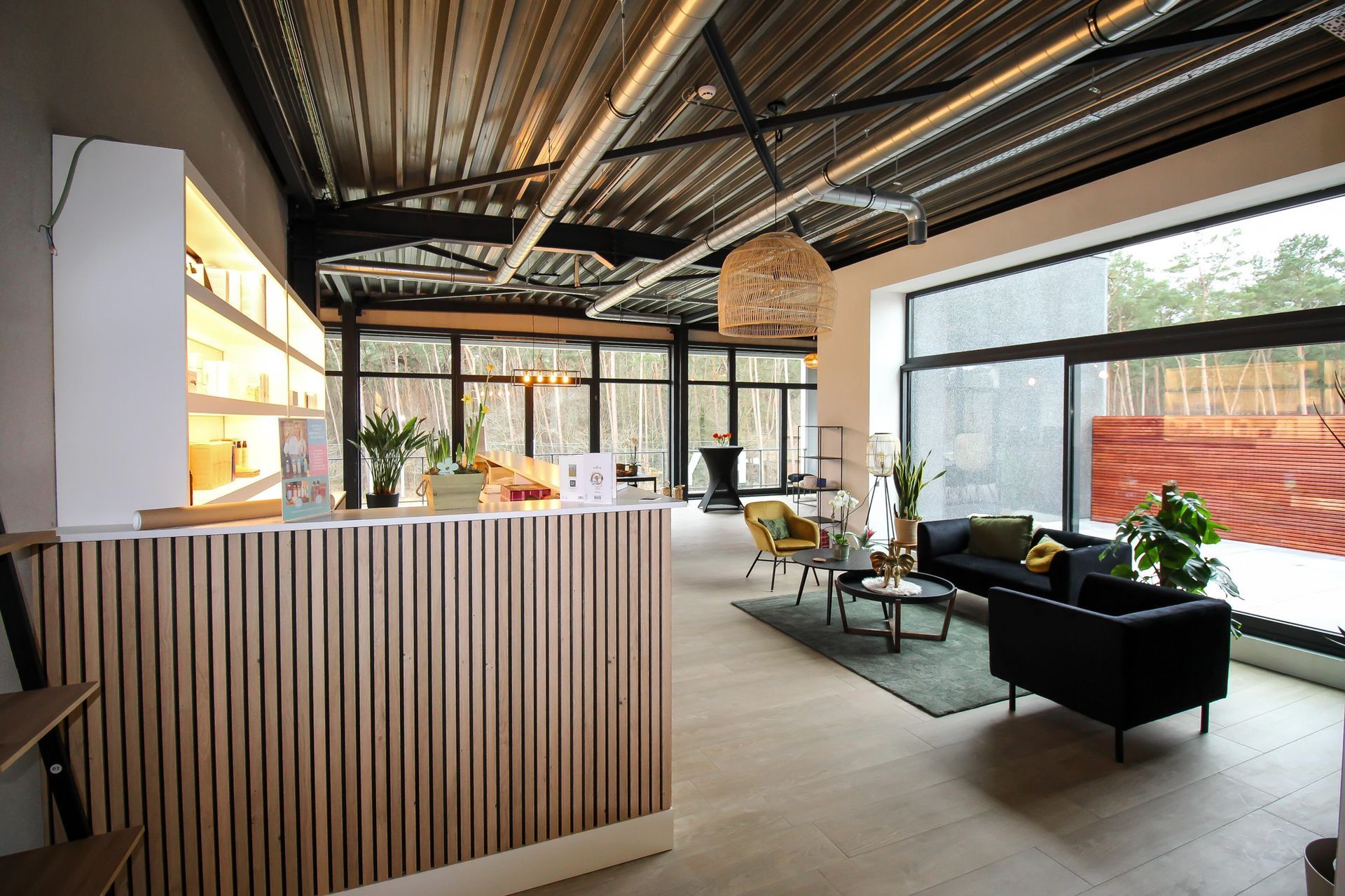 Ontvangstruimte met houten balie en wachtruimte met bank en stoelen; grote ramen met uitzicht op het bos.