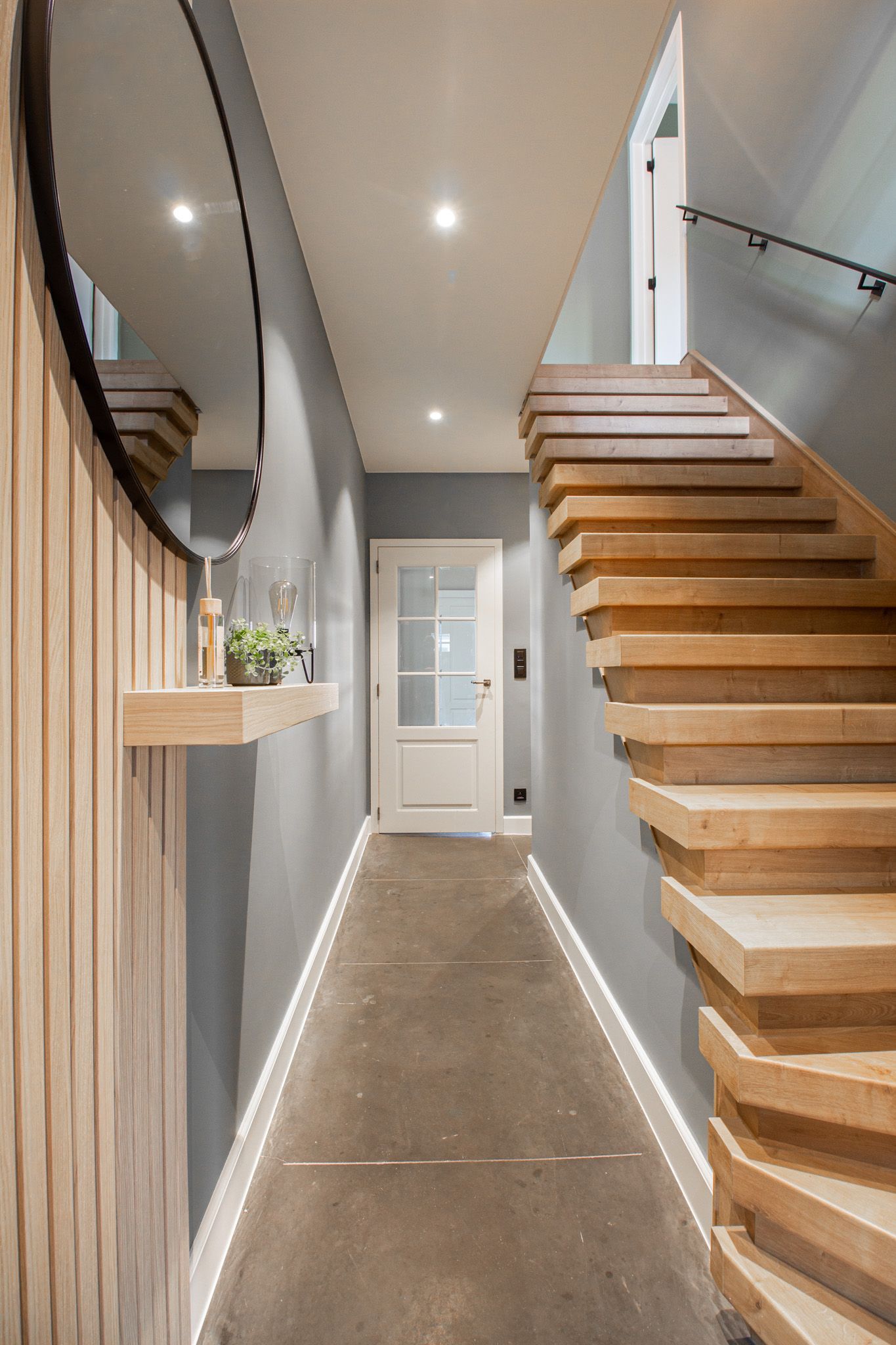 Smalle gang met houten trap en lambrisering, grijze muren en een witte deur aan het einde.