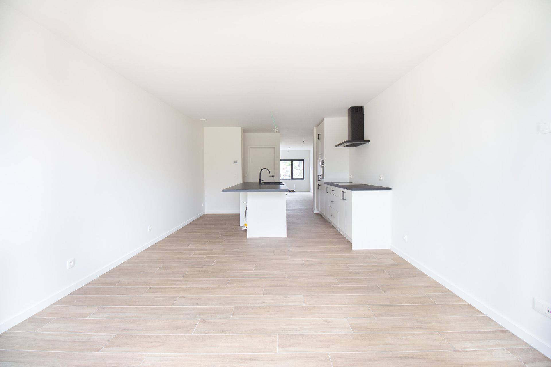 Lege, lichte keuken met witte kasten, zwart aanrechtblad en lichte houten vloer.