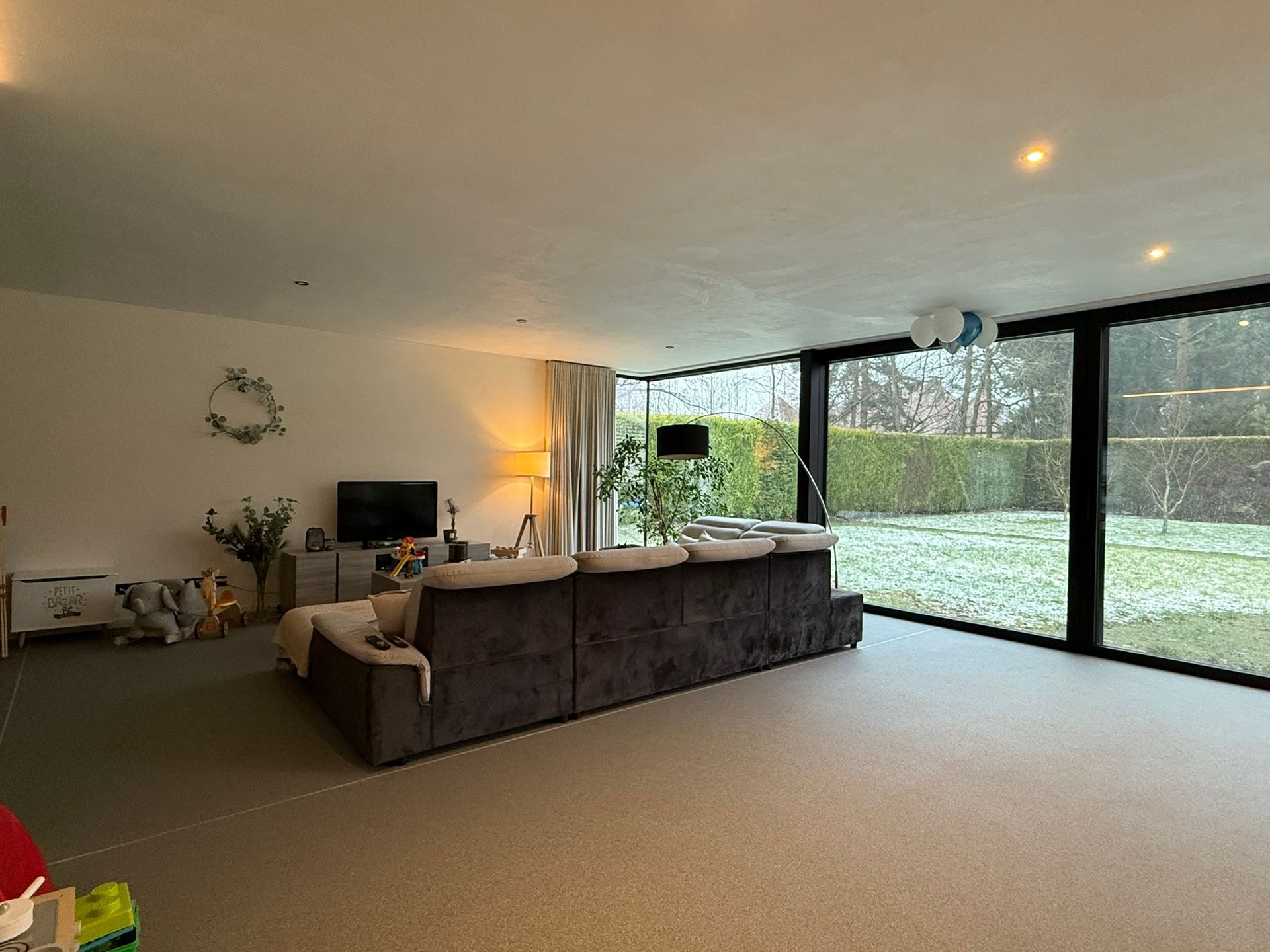 Ruime woonkamer met donkere bank, grote ramen met uitzicht op de achtertuin en tv.