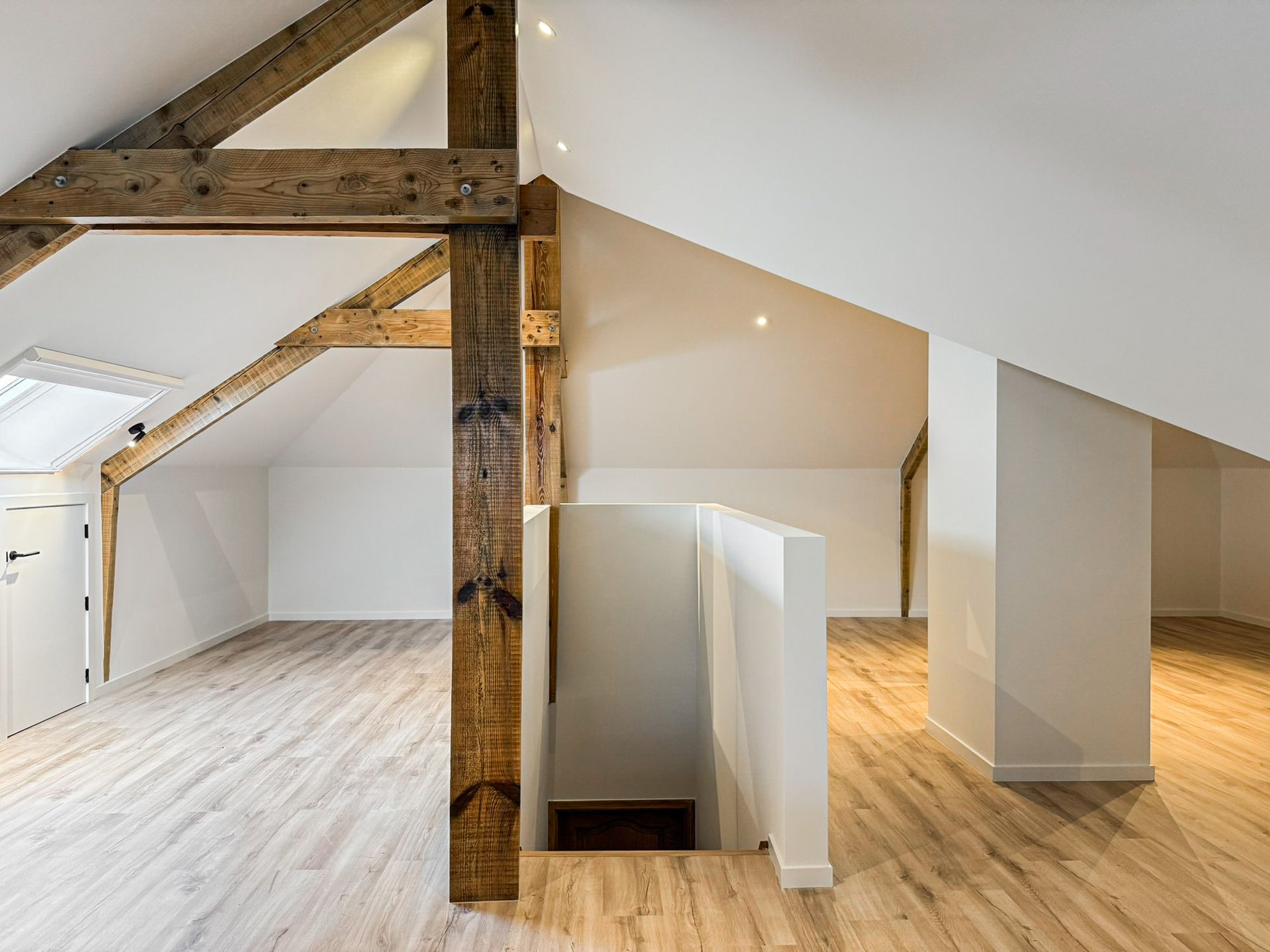Lichte zolderruimte met zichtbare houten balken, witte muren en een lichte houten vloer.