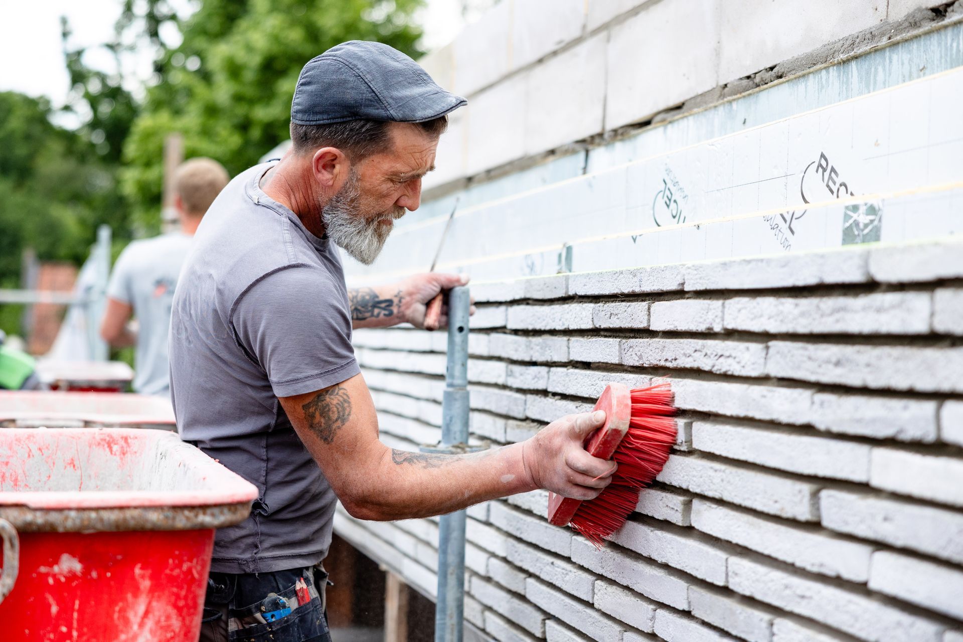 A construction worker spreading mortar with a trowel on a brick wall. Scaffolding and red bucket in view.