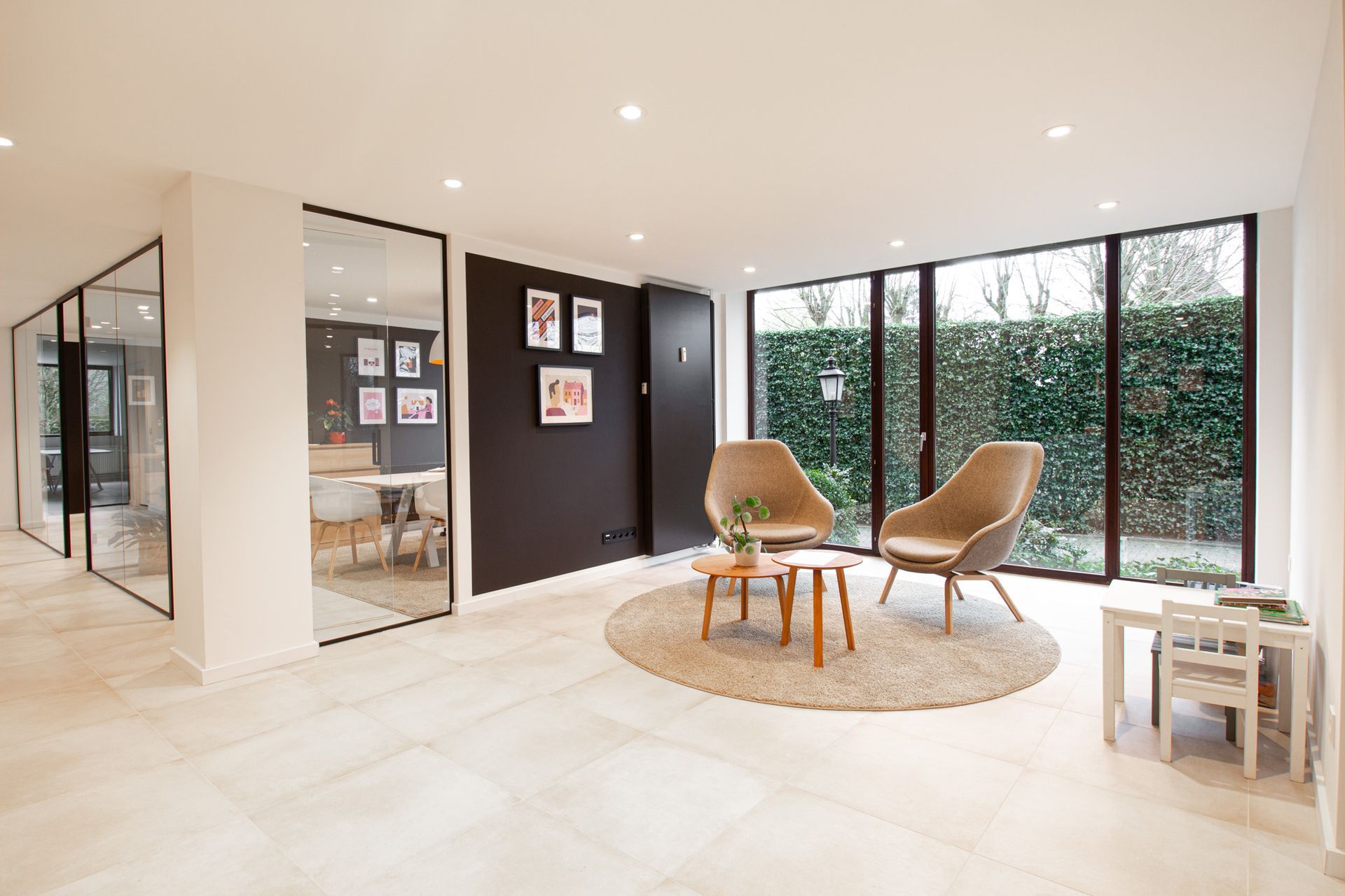 Moderne wachtkamer met bruine accentmuur, glazen wanden, zithoek en klein tafeltje, met uitzicht op een heg.