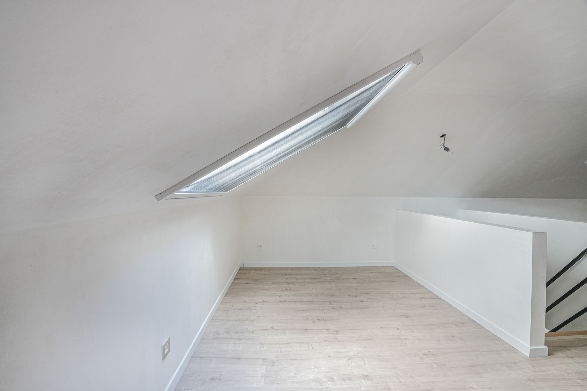 Lege zolderkamer met schuin plafond, metalen ventilatieopening en witte muren. Lichte houten vloer en witte balustrade.