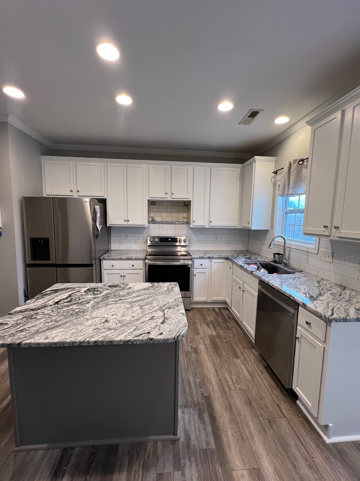Modern kitchen with white cabinets, granite countertops, a dark gray island, stainless steel appliances, and wood floors.