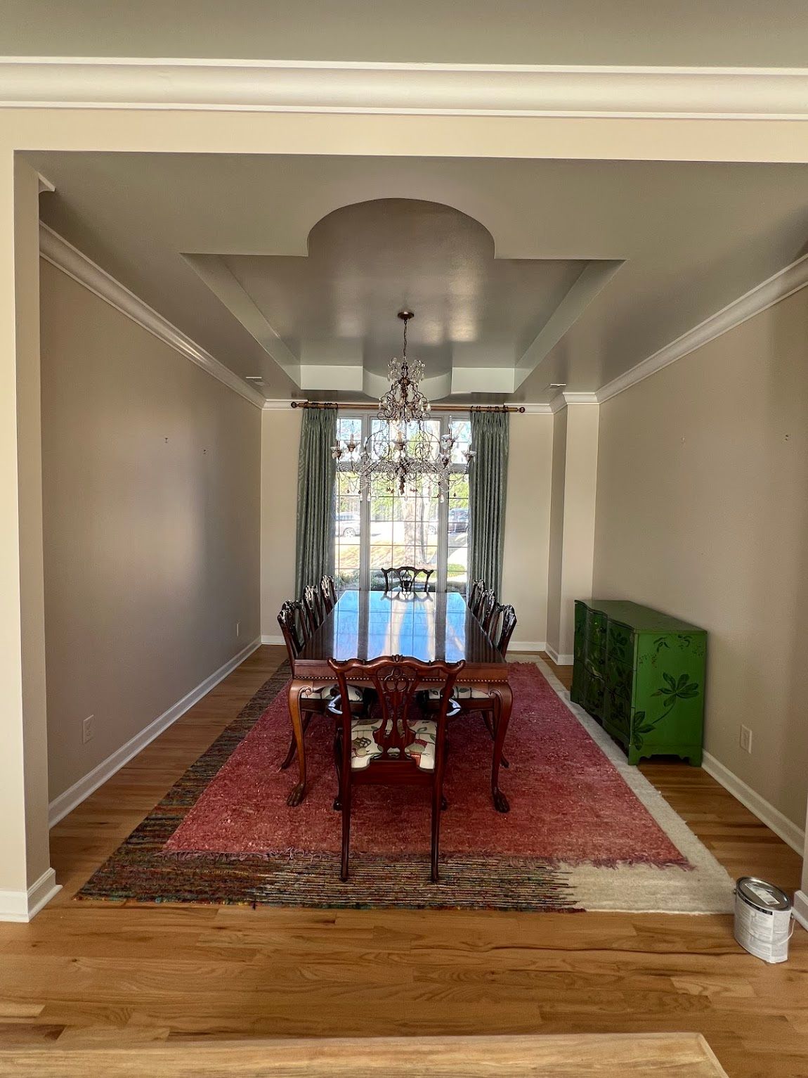A dining room with a long table, chairs on a red patterned rug, a green cabinet, and a chandelier under a tray ceiling.