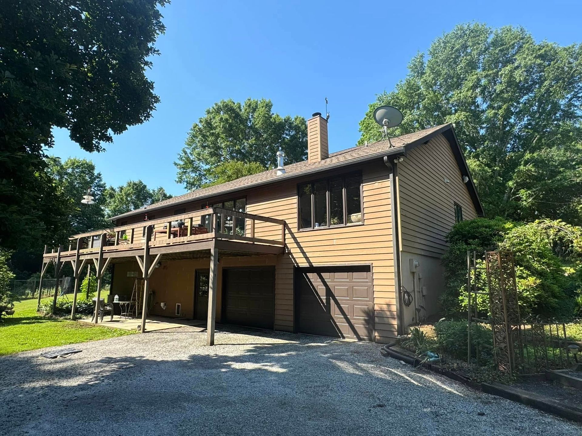 A two-story, brown wood-sided house with a large upper deck and a two-car garage, set on a gravel driveway amid trees.