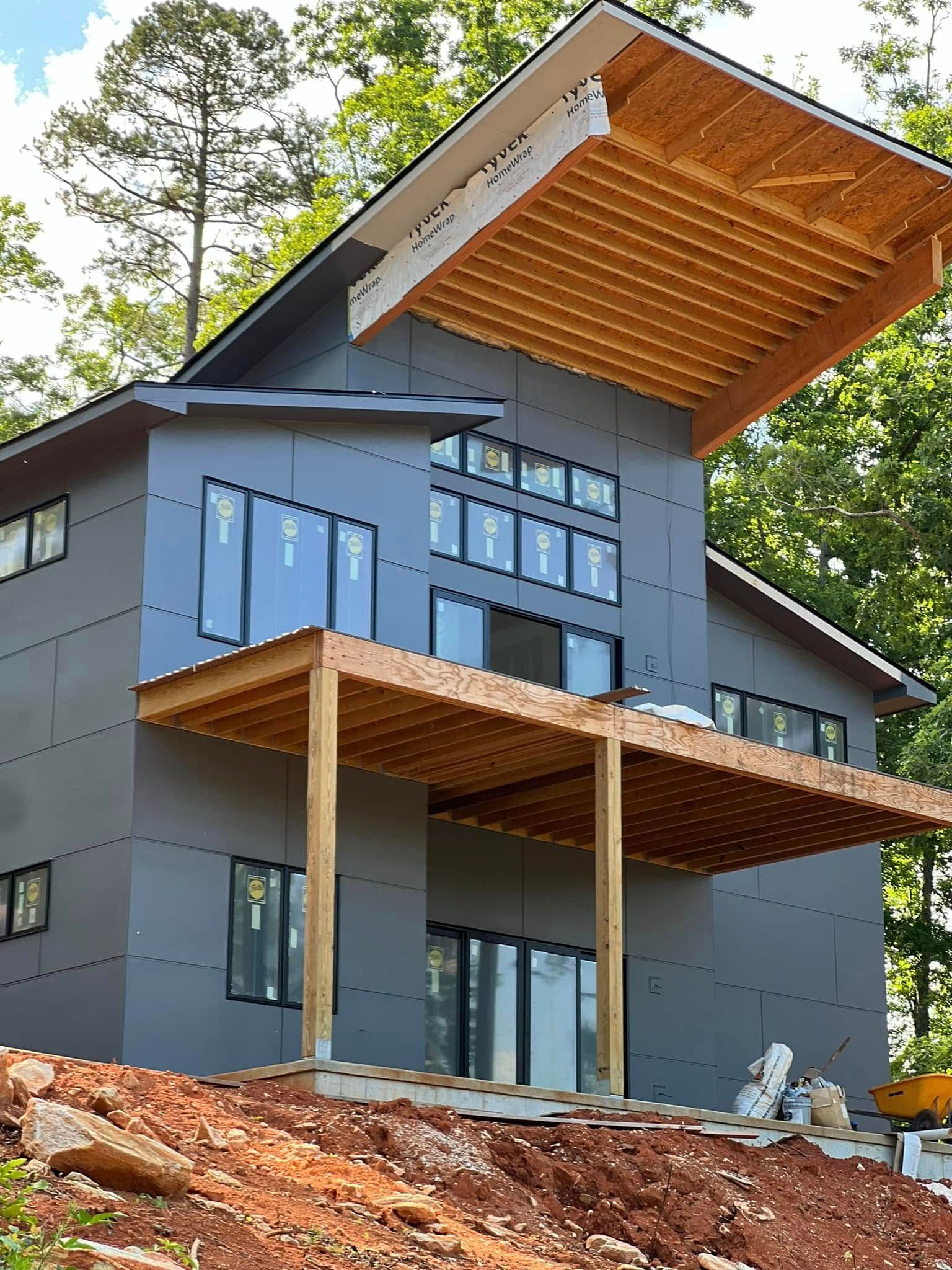 A house under construction with grey exterior sheathing, a large overhanging wooden roof deck, and a wooden porch.