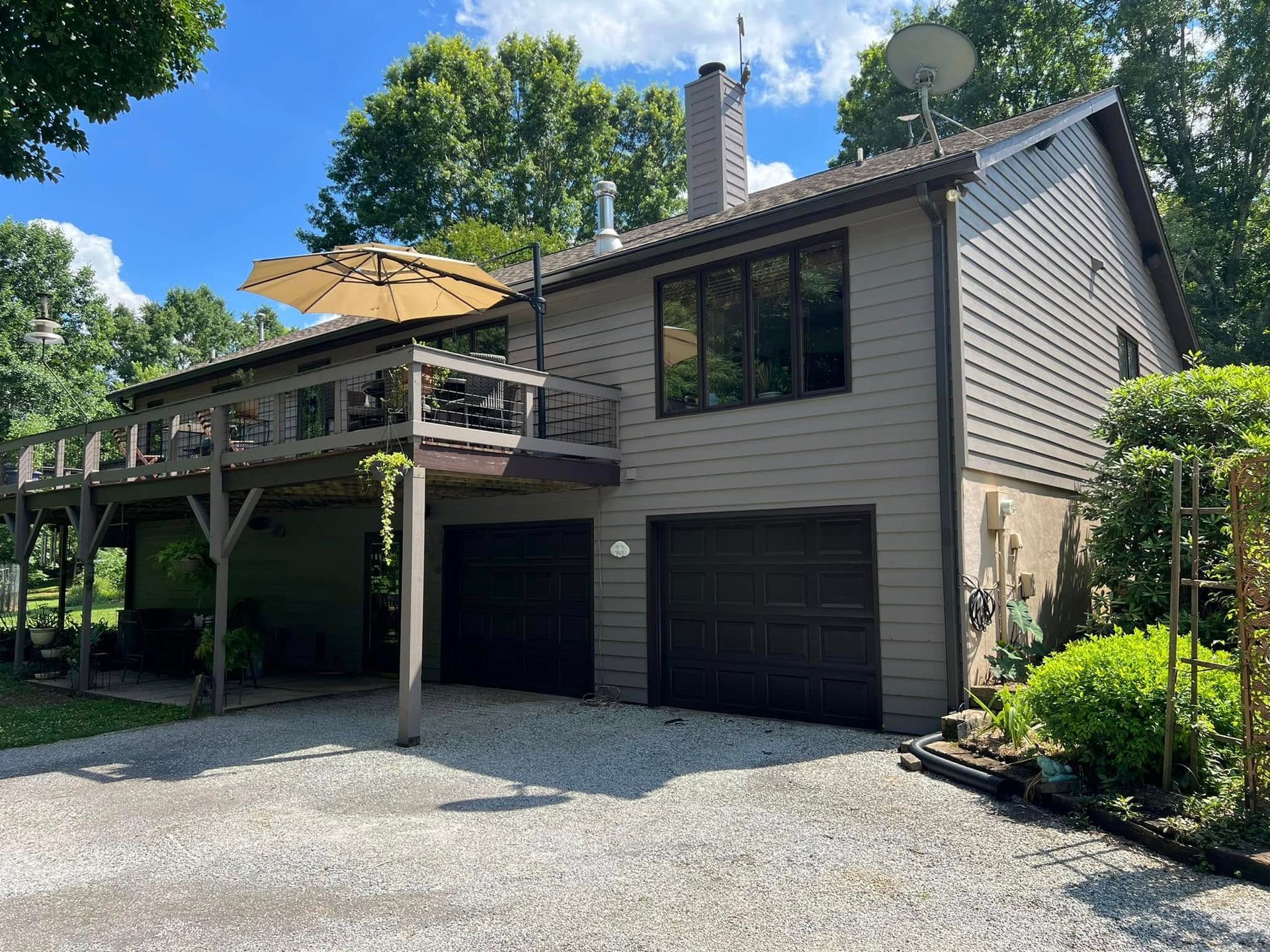 A two-story tan house with a gravel driveway, a large raised deck, a beige umbrella, and a two-car garage.