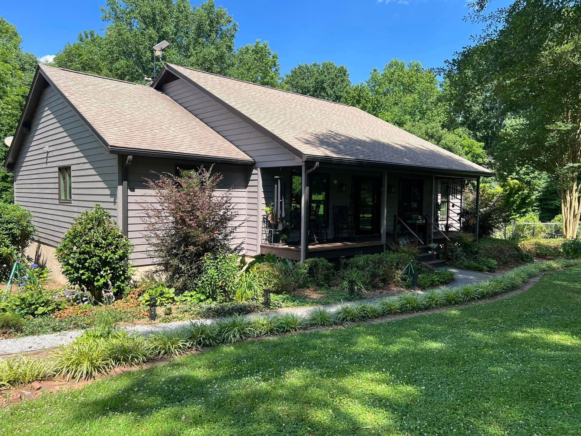 A brown house with a wrap-around porch, nestled in a lush, green, sunlit yard surrounded by dense trees.