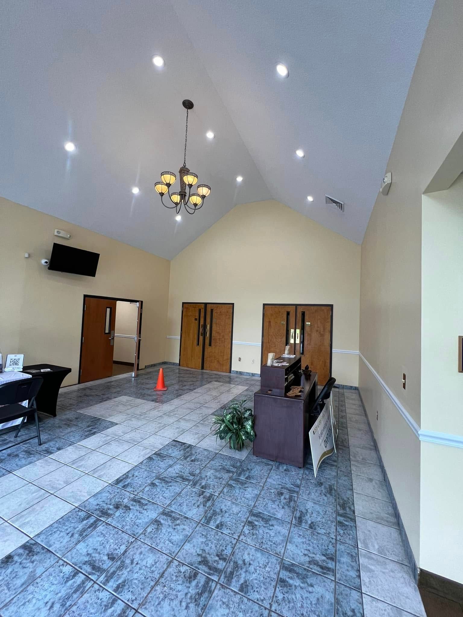 A vaulted-ceiling hall with patterned blue carpet, light walls, wooden double doors, a chandelier, and a front desk.